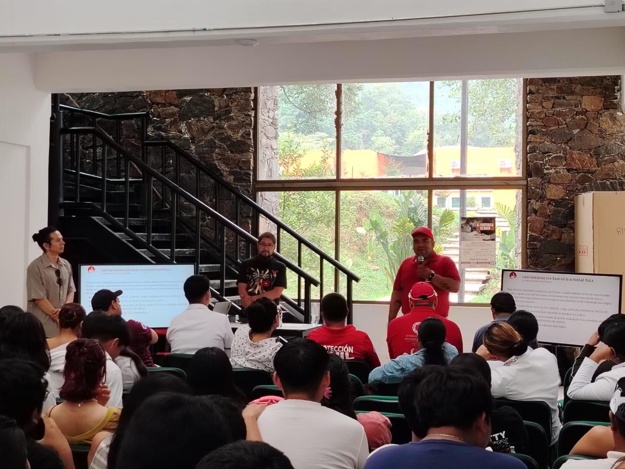 Speaker in red addresses seated audience in a brick-walled room with stairs and a whiteboard.