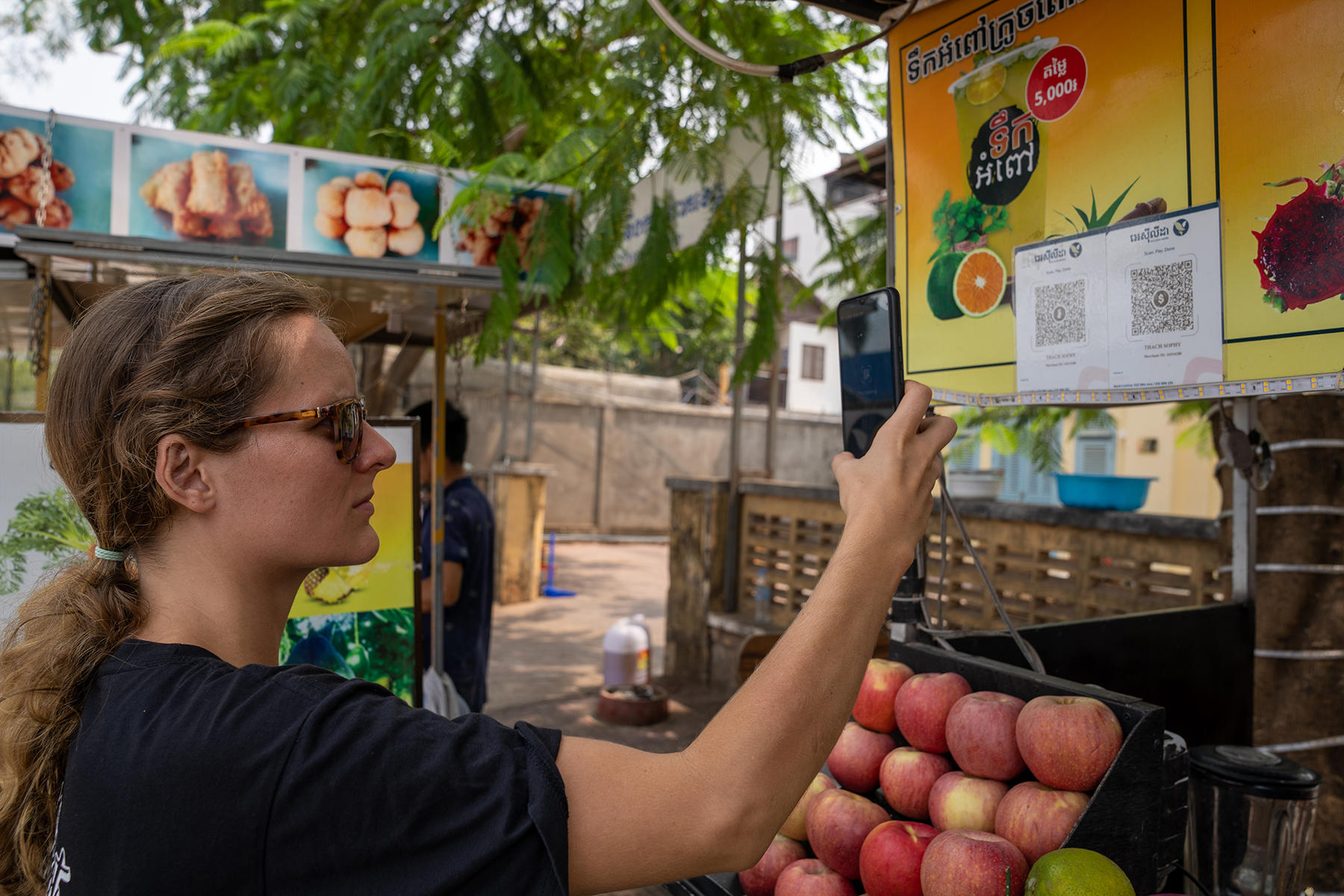 Woman with ponytail checking a QR code with her phone, at an outdoor market near crates of apples.