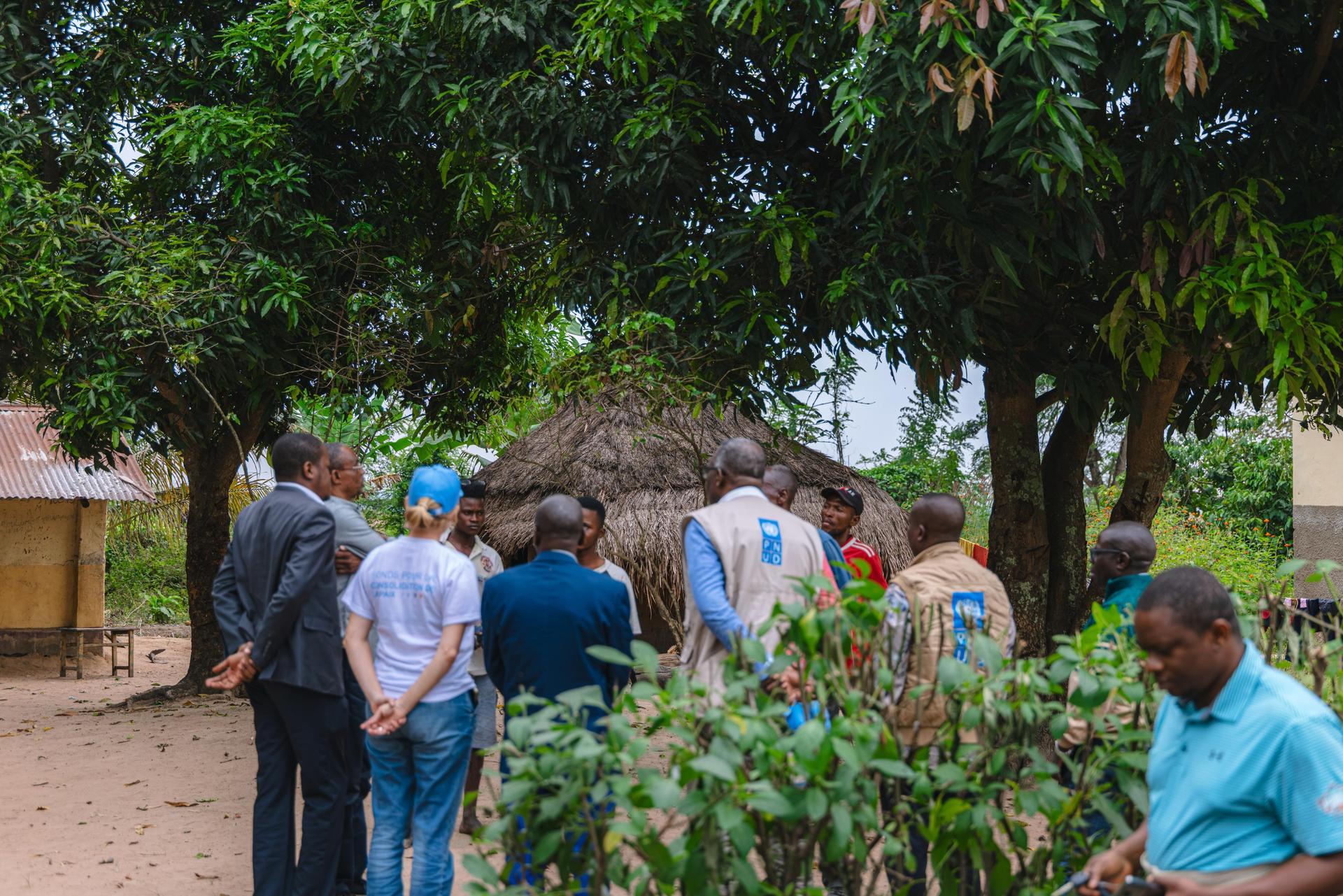 Group of people standing in a village yard with thatched huts and trees.