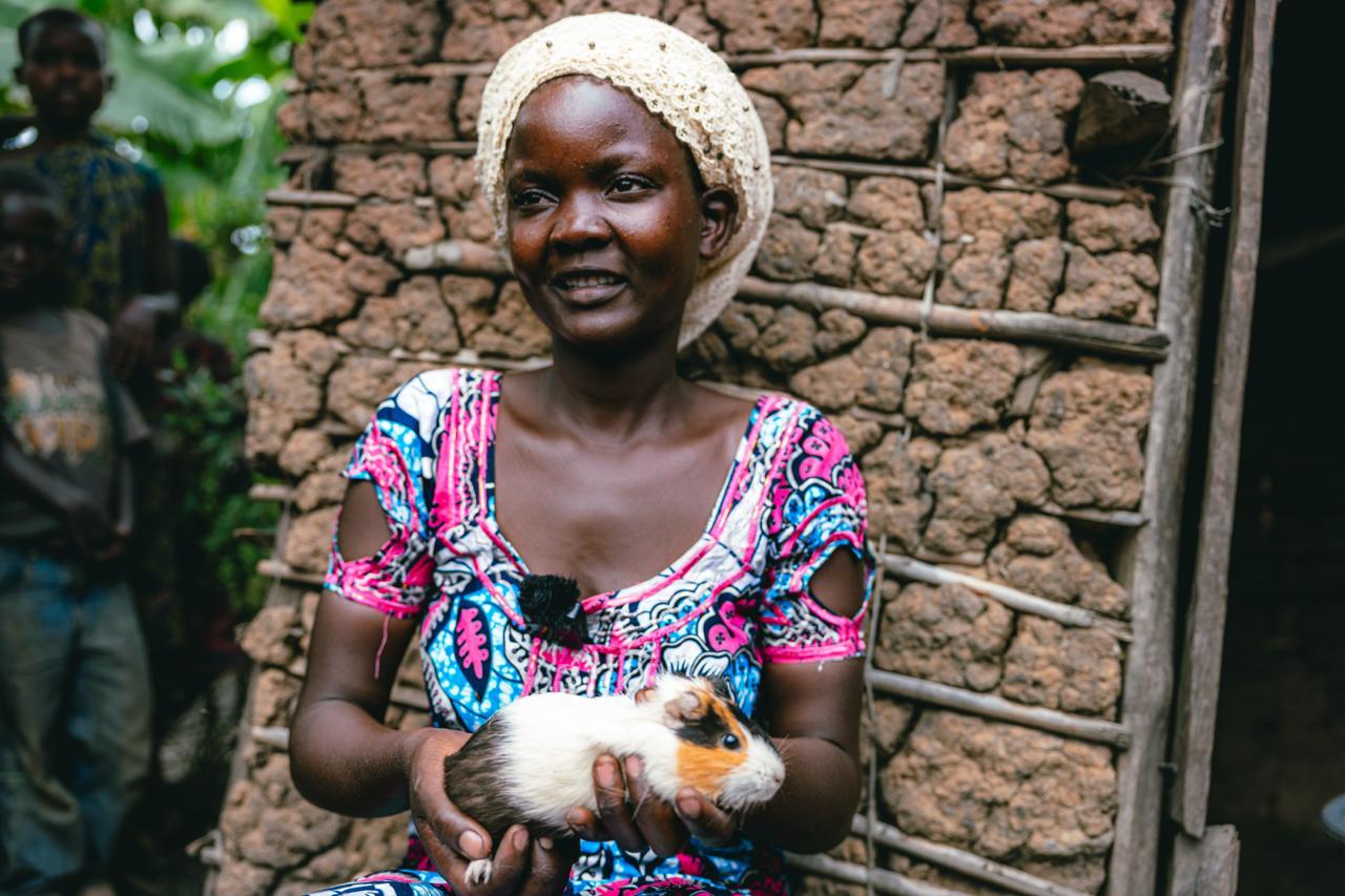 Woman with a blurred face in a colorful dress holds a chick beside a mud-brick wall.