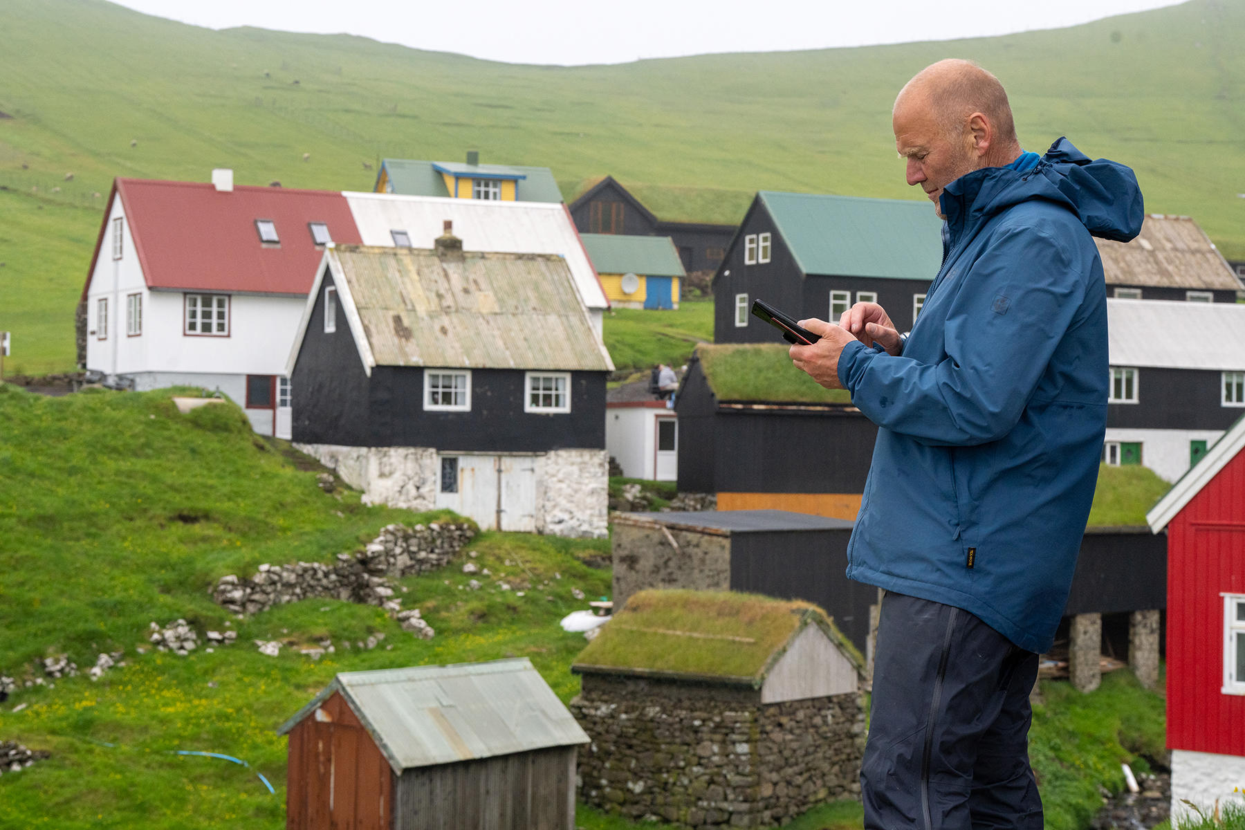 Man in a blue jacket on a grassy hill, holding a phone, overlooking a cluster of colorful houses.