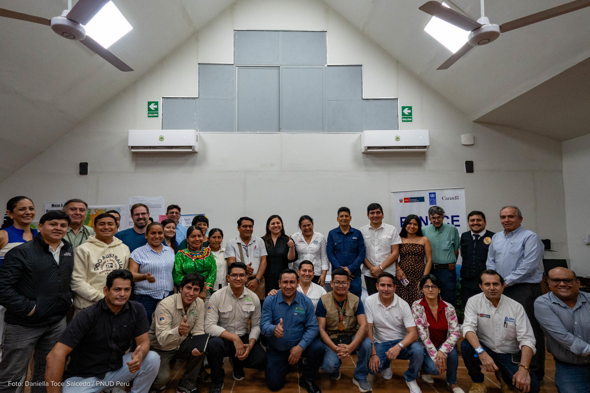 Large group of people posing for a group photo in a meeting room with ceiling fans.