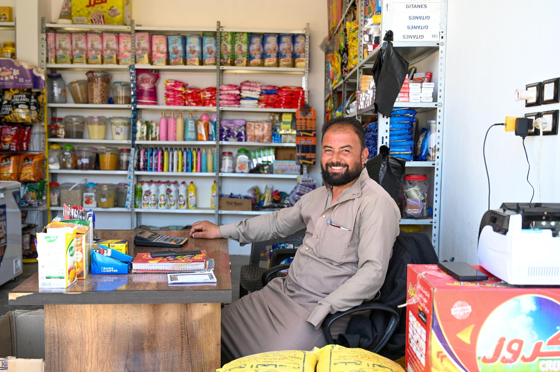 Photo of a man seated at a small shop counter with shelves full of snacks behind him.