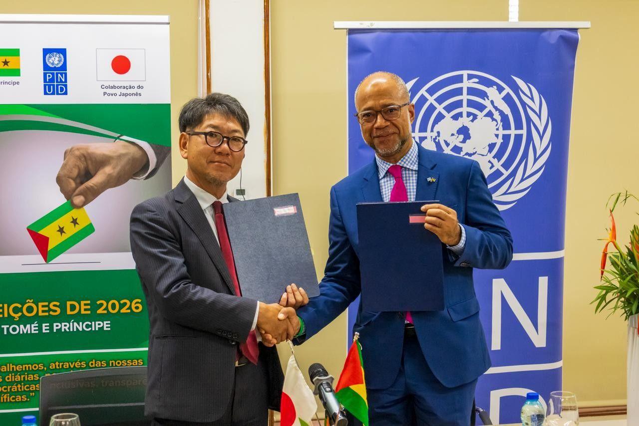 Two men in suits shake hands, each holding a document; UN banner and election poster in background.