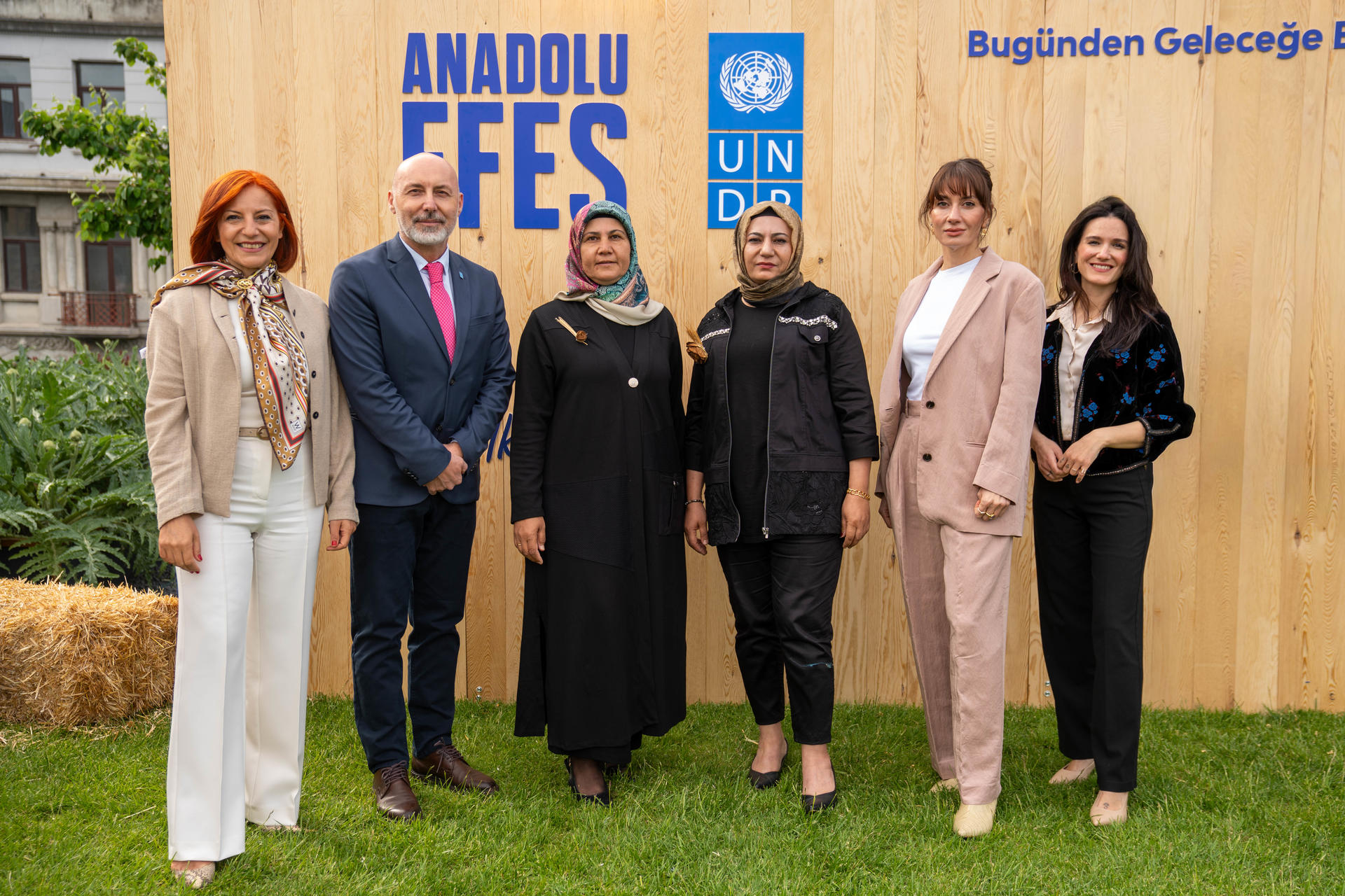 Seven people pose outdoors in front of a wooden wall with Anadolu signage and UN logos.
