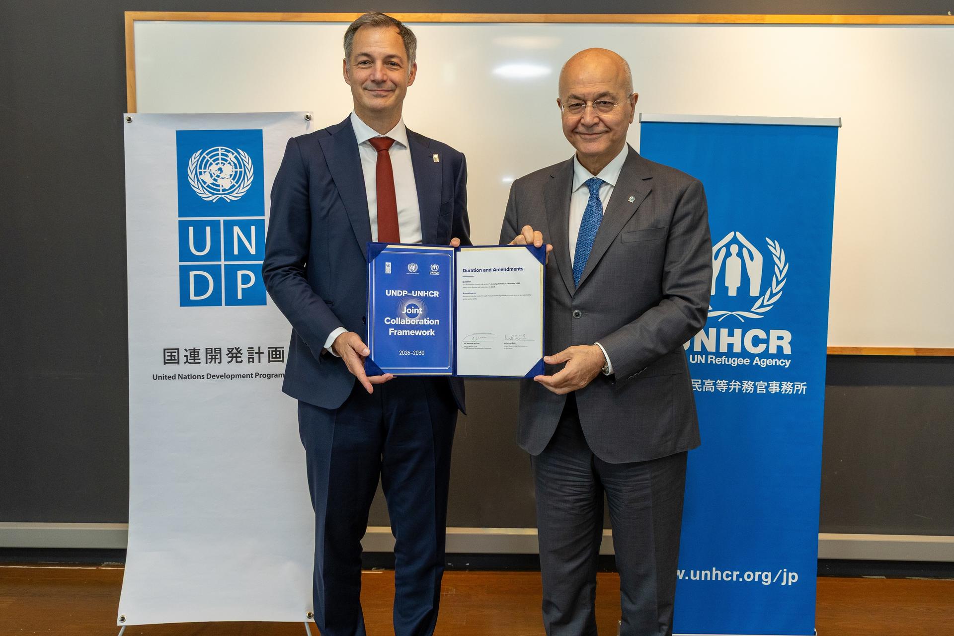 Two men in suits smile while holding a certificate beside UNDP and UNHCR banners.