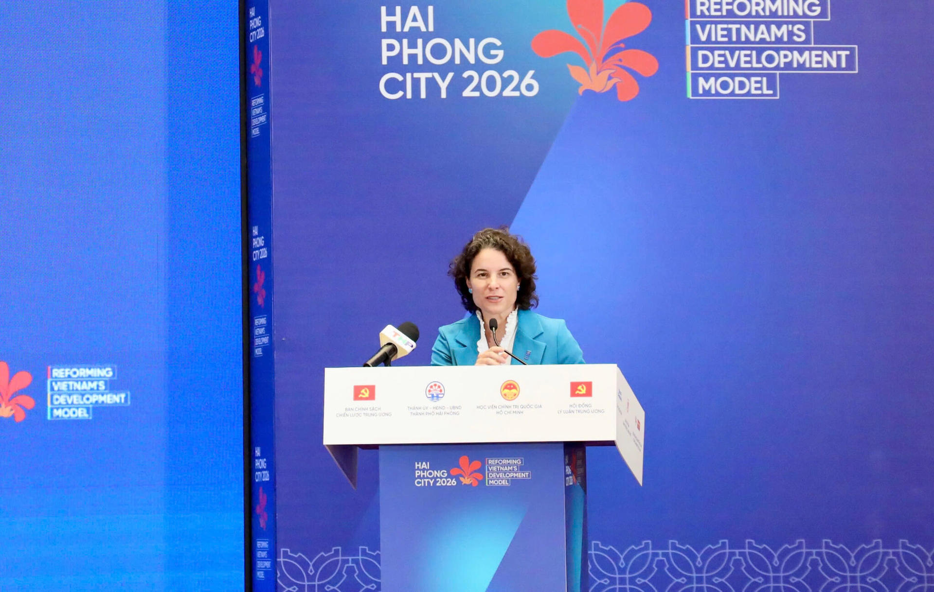 Female speaker at a podium on a blue conference stage with banners.