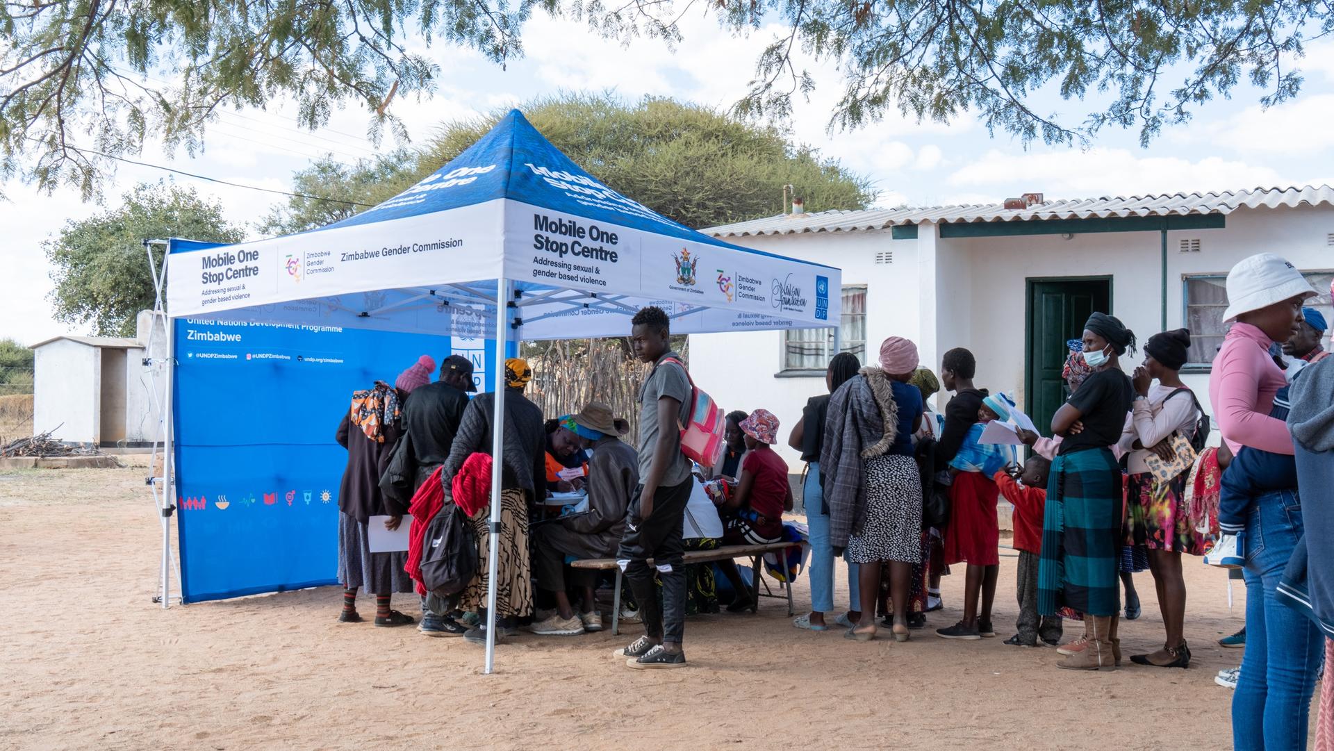 Photograph: People queue under a blue tent outside a white building.