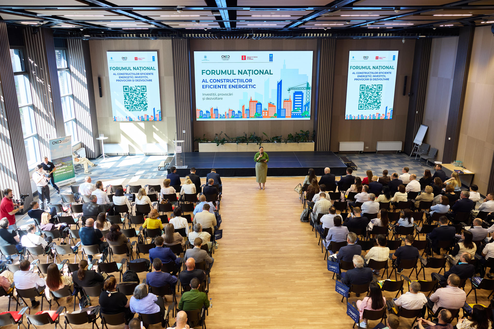 Photograph of a large auditorium with an audience watching a speaker at a podium and two screens.