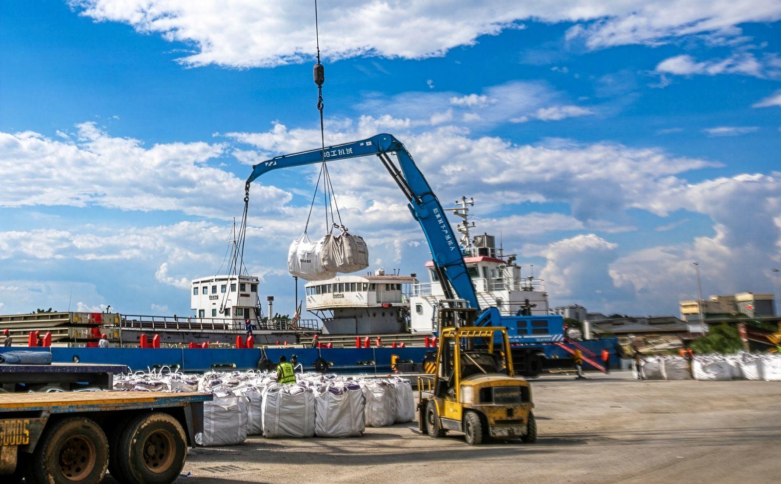 Photograph of a harbor construction site with a blue crane lifting a load; ships in background.