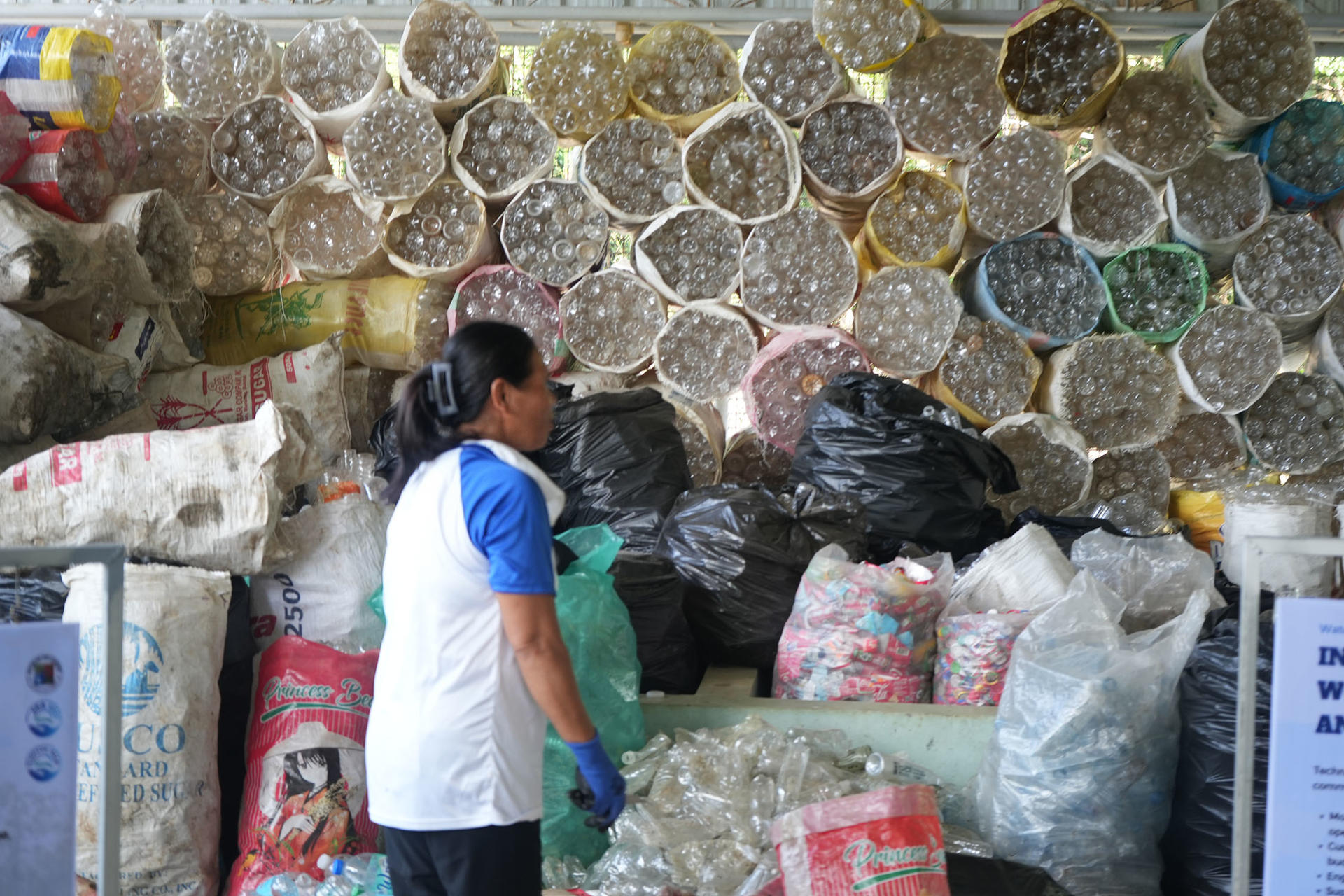 Man in blue and white shirt stands near stacked circular bales and bags of recyclables.