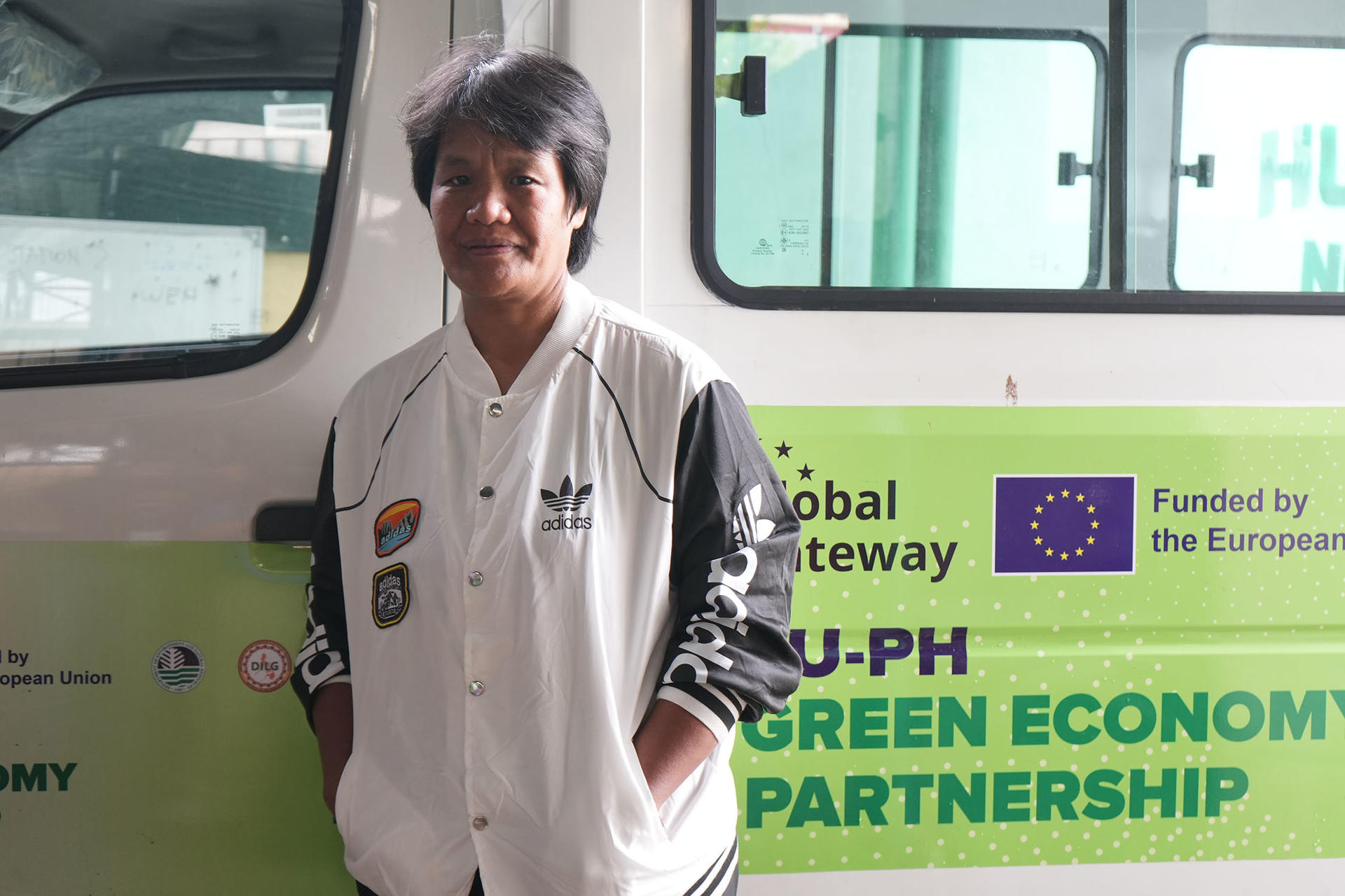 Person in a white lab coat stands beside a green-and-white bus branded with Green Economy Partnership