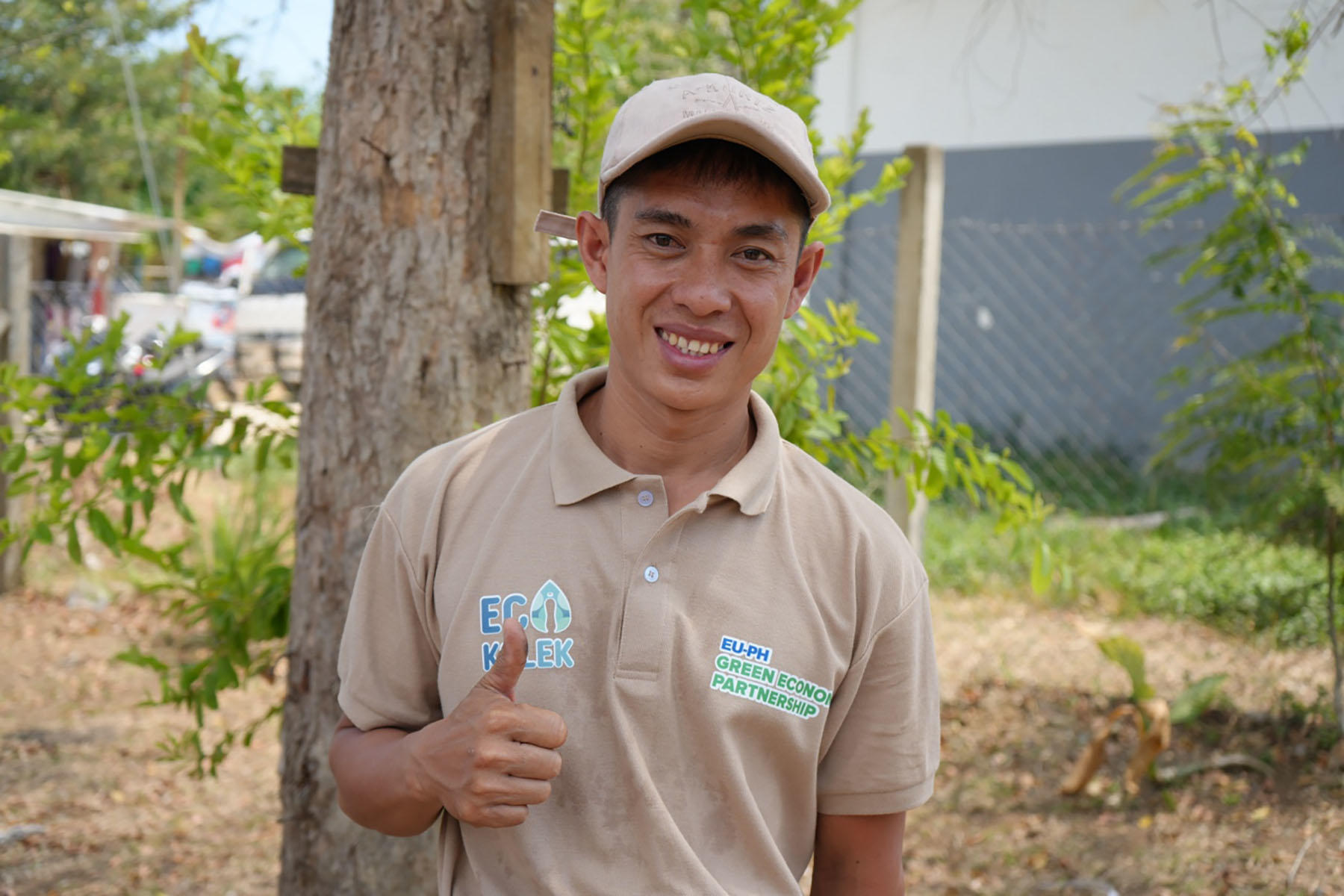 Beige polo-clad man with blurred face giving thumbs up outdoors, tree and building in background.
