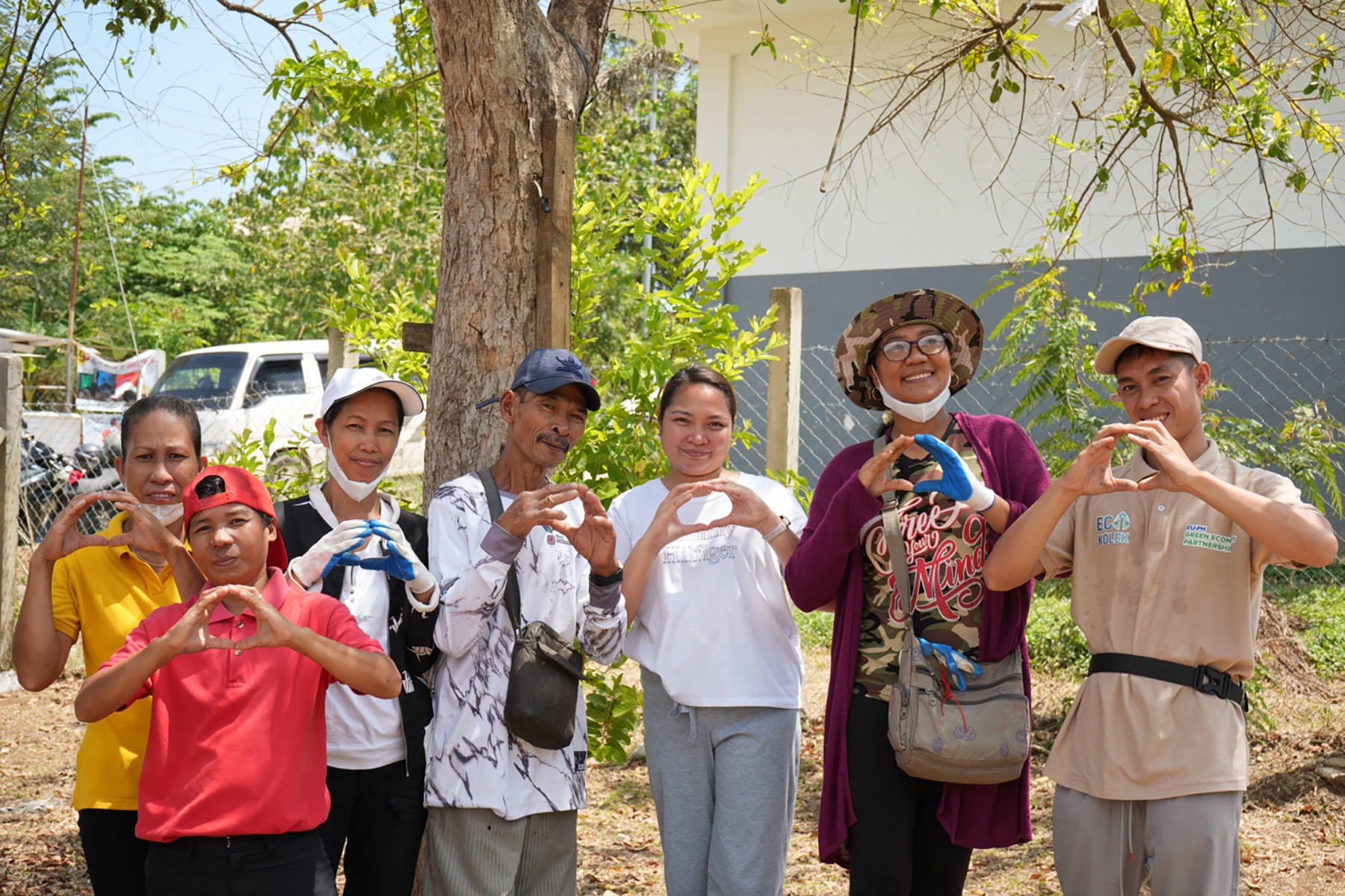 Group of diverse adults posing outdoors, smiling; trees and cars visible in the background.