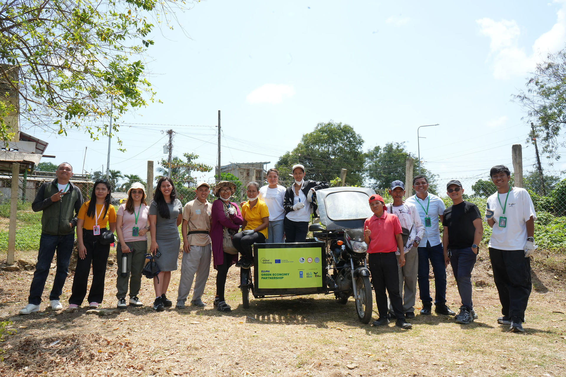 Photograph of a diverse group of people standing in a line outdoors on a sunny day around a yellow signboard on a cart.