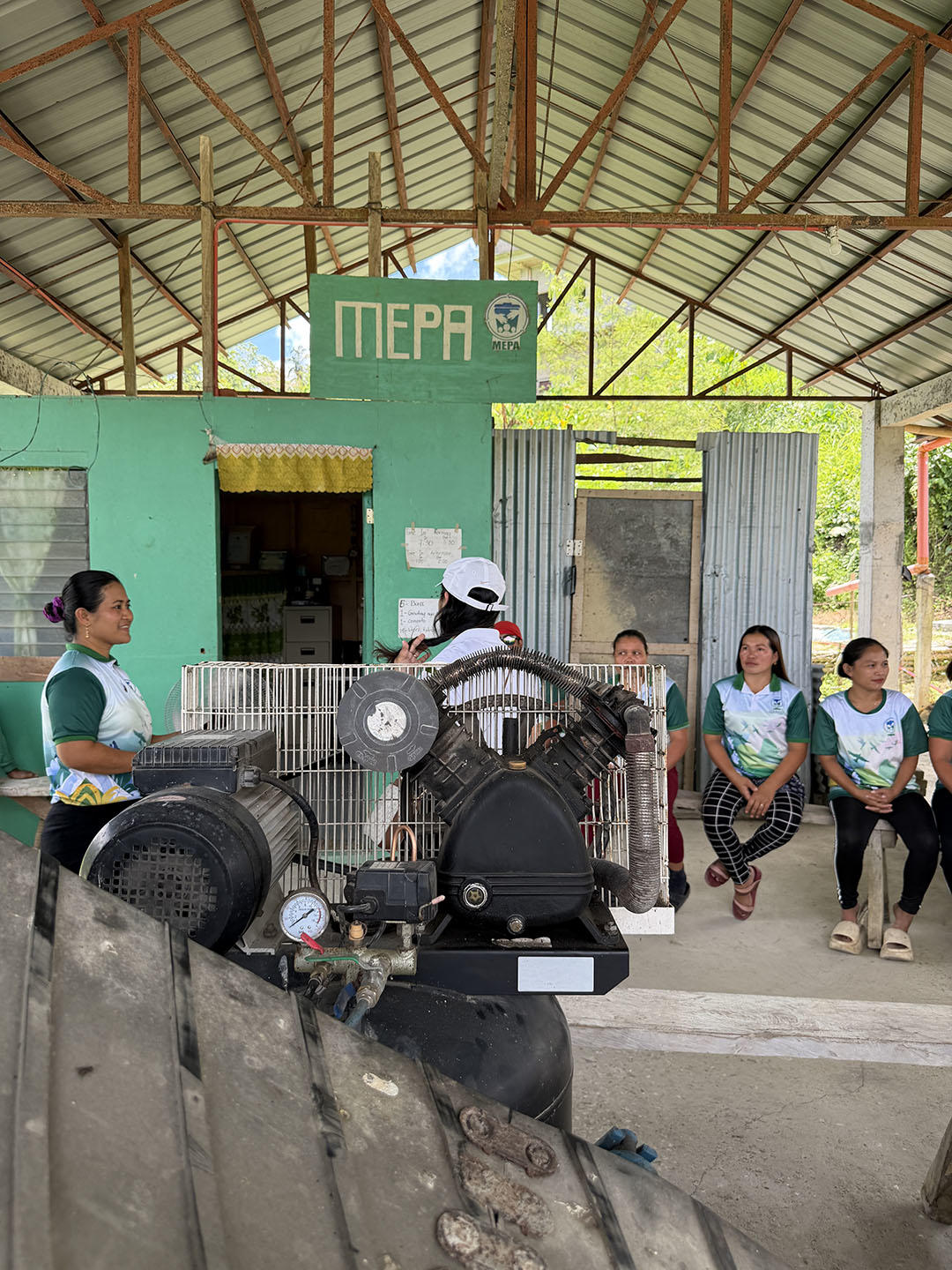 Photo: group of people outside green building under a metal roof; a man in a helmet near machinery