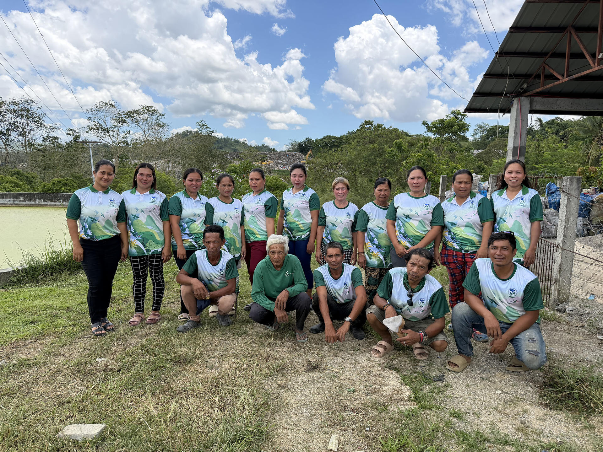 Group of people in teal shirts posing in two rows on a sunny outdoor field, shelter to the right.