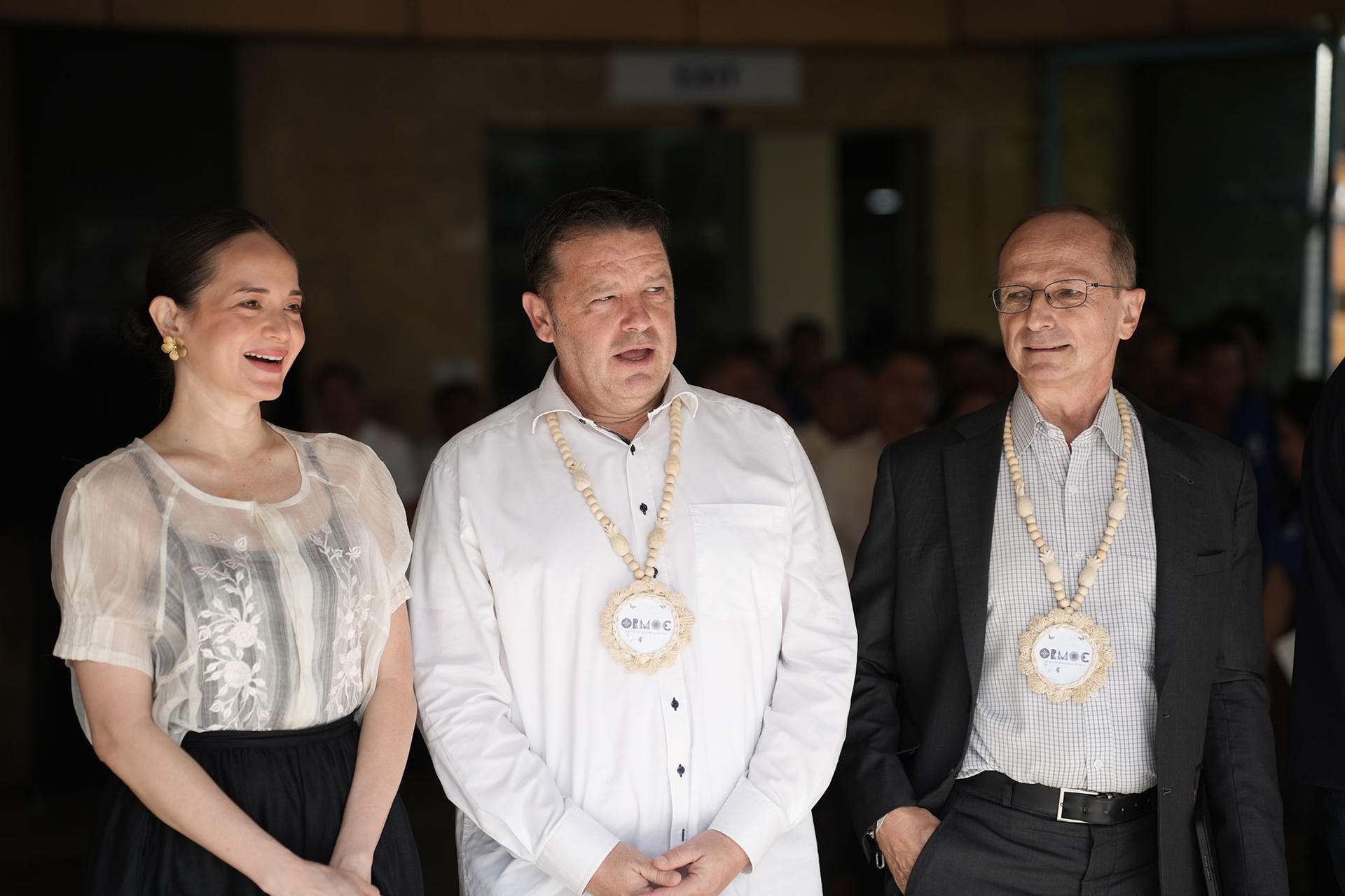 Three formally dressed people with ceremonial sashes and medallions pose indoors; faces blurred.