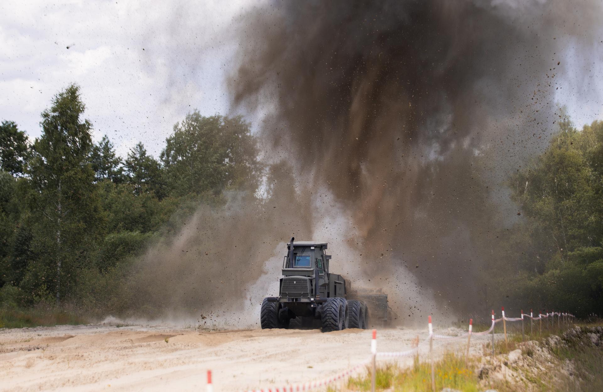 Photograph of a blue tractor on a dusty dirt road, large dust cloud rising behind.