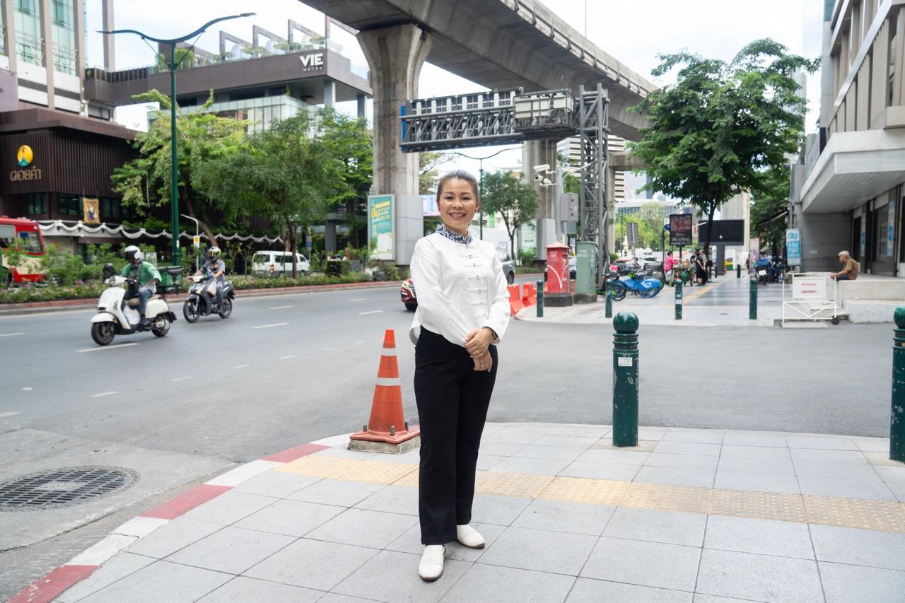 Smiling woman in white blouse and black pants stands on a city sidewalk under an elevated road.