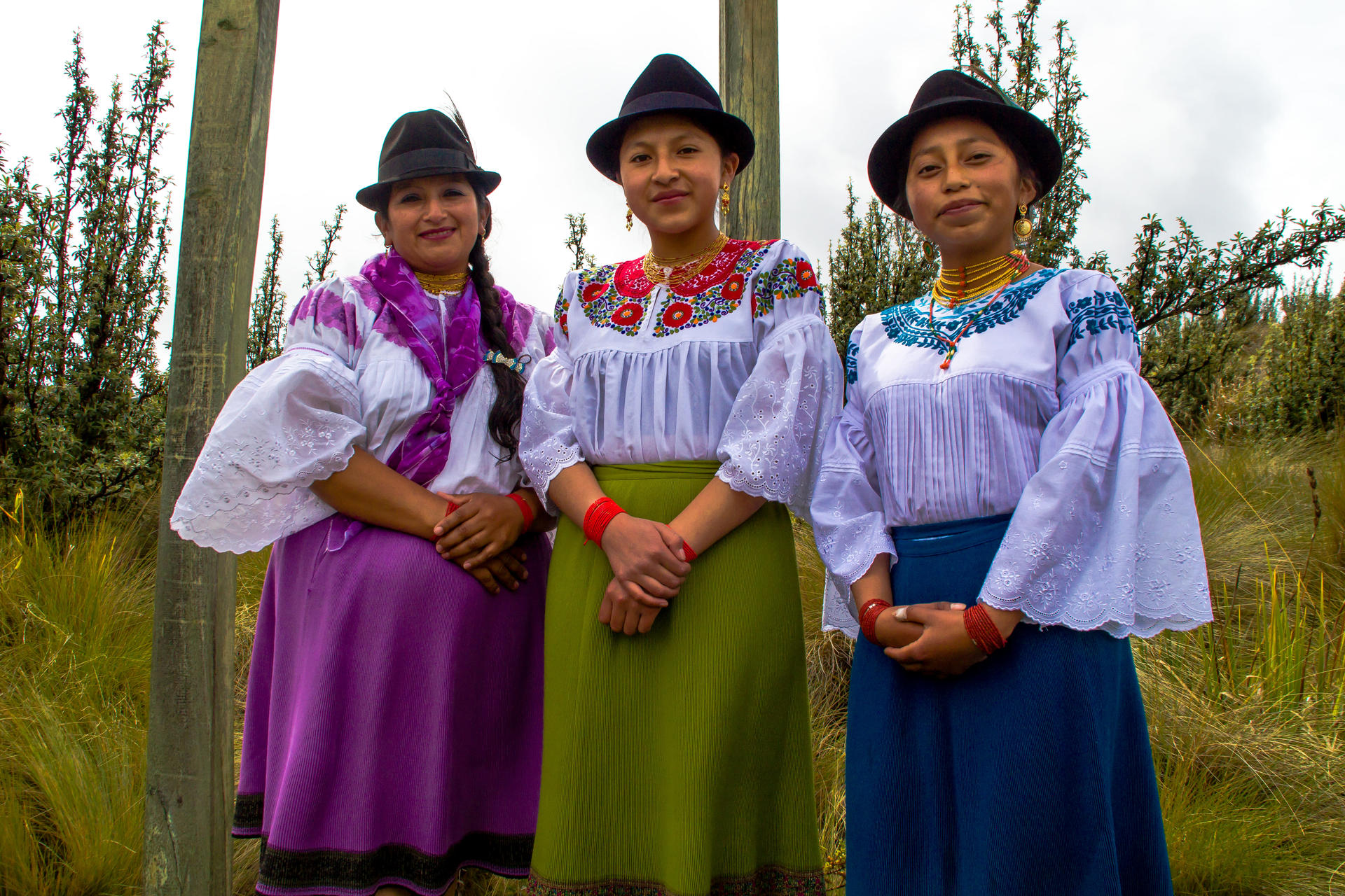 Three people in colorful traditional dresses and hats pose in a grassy field.