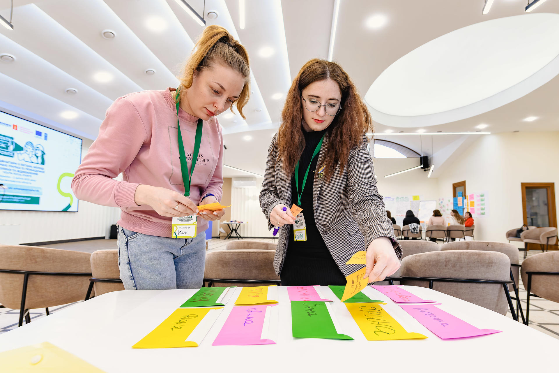Two women with lanyards stand at a table covered in colorful sticky notes in a bright conference room.