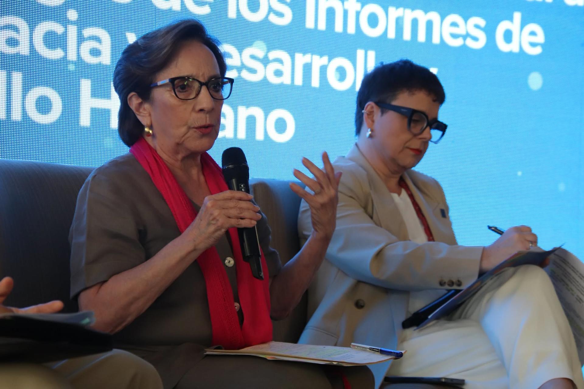 Two panelists on stage with microphones, blurred faces, seated before a blue backdrop.