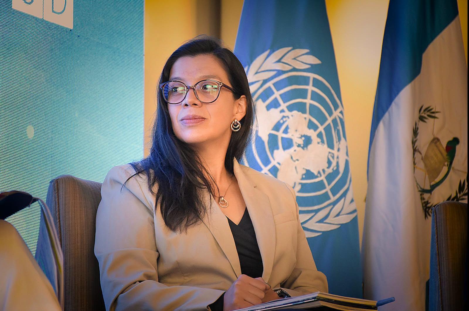 Photo of a blurred woman in a beige blazer at a desk, with UN flags and emblem in the background.