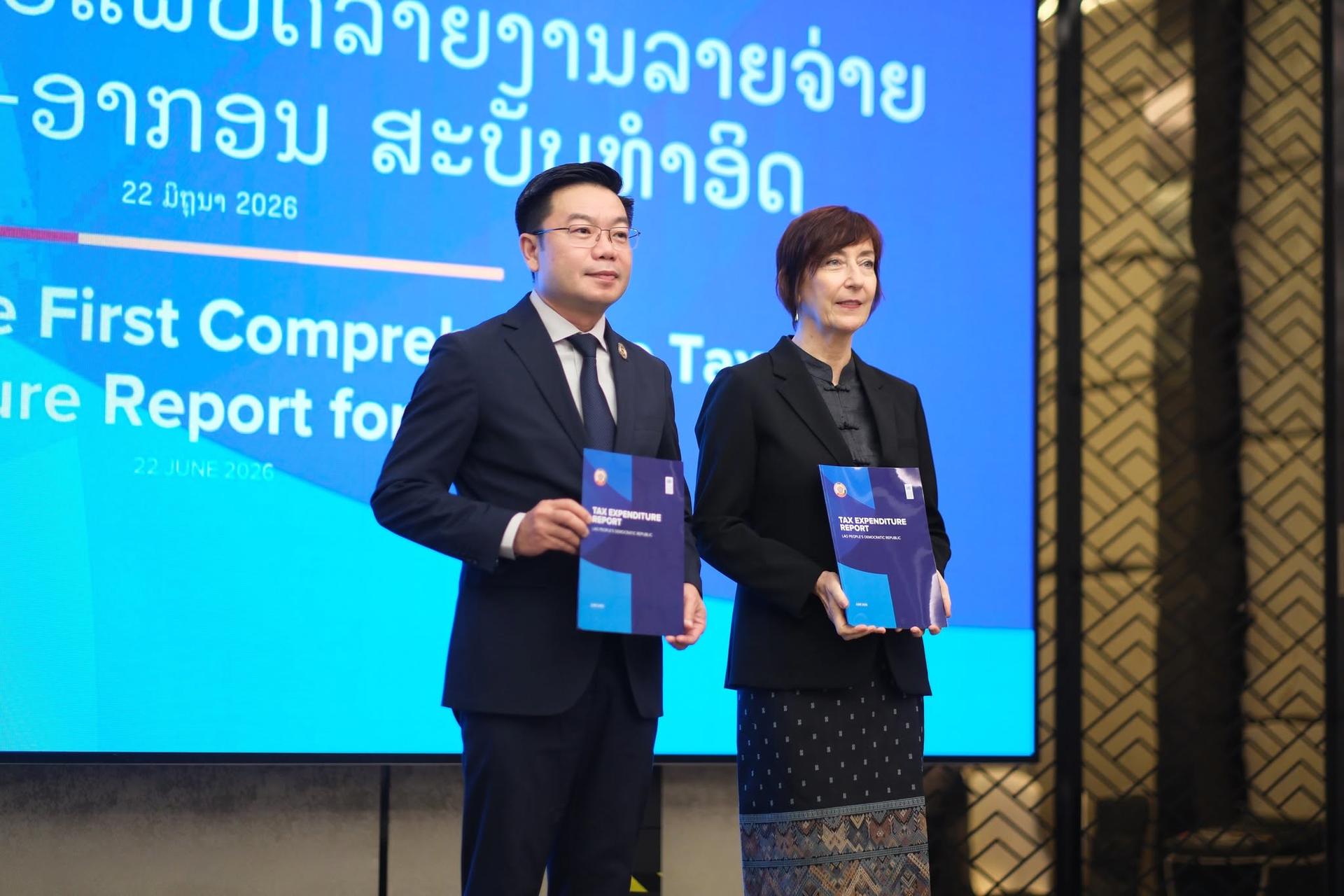 Two formally dressed individuals on stage, holding blue folders, with a blue display behind.