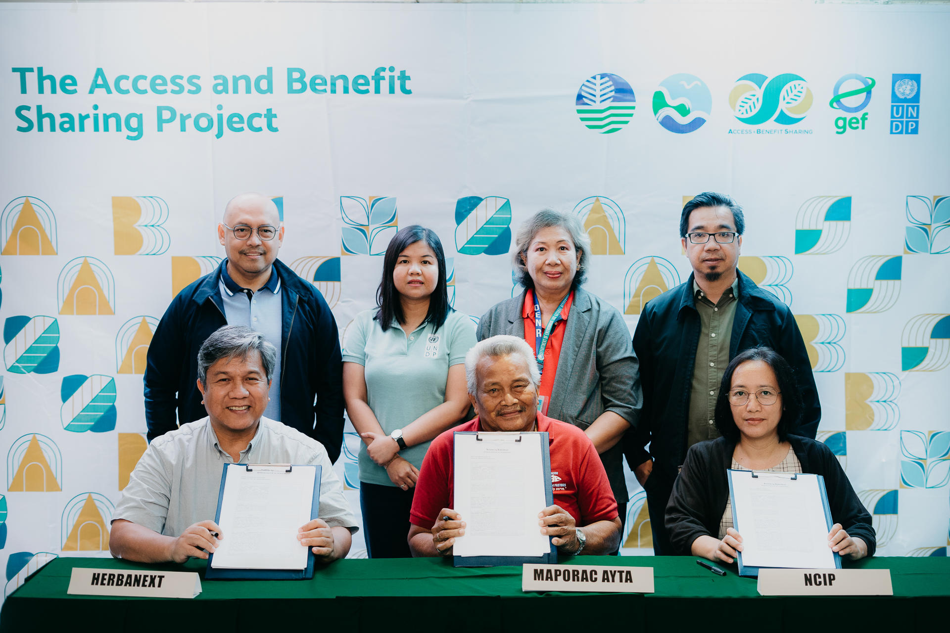 Photograph of a signing ceremony with three people seated in front holding documents.