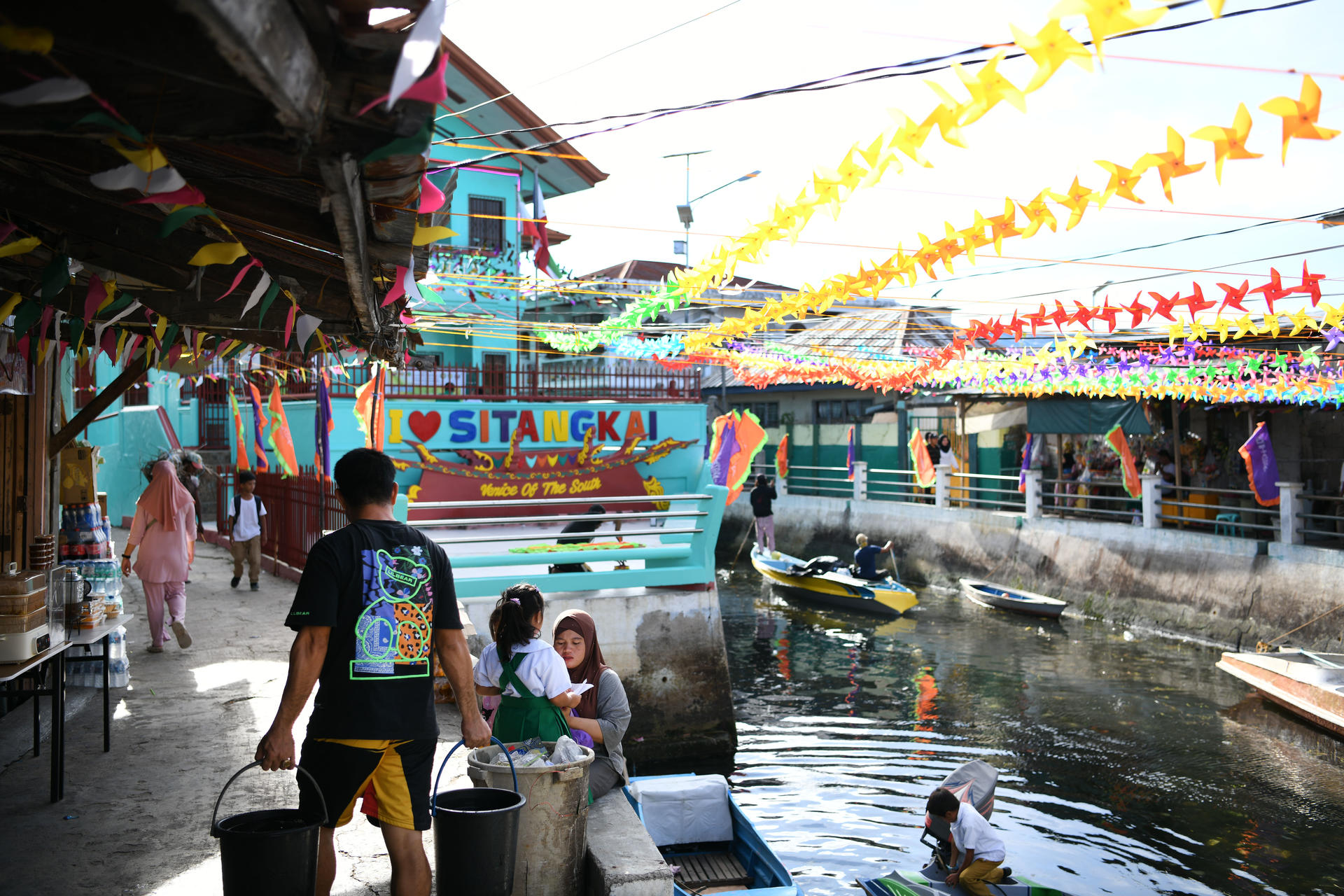 People at a floating market along a canal, with colorful pennant banners and boats nearby.