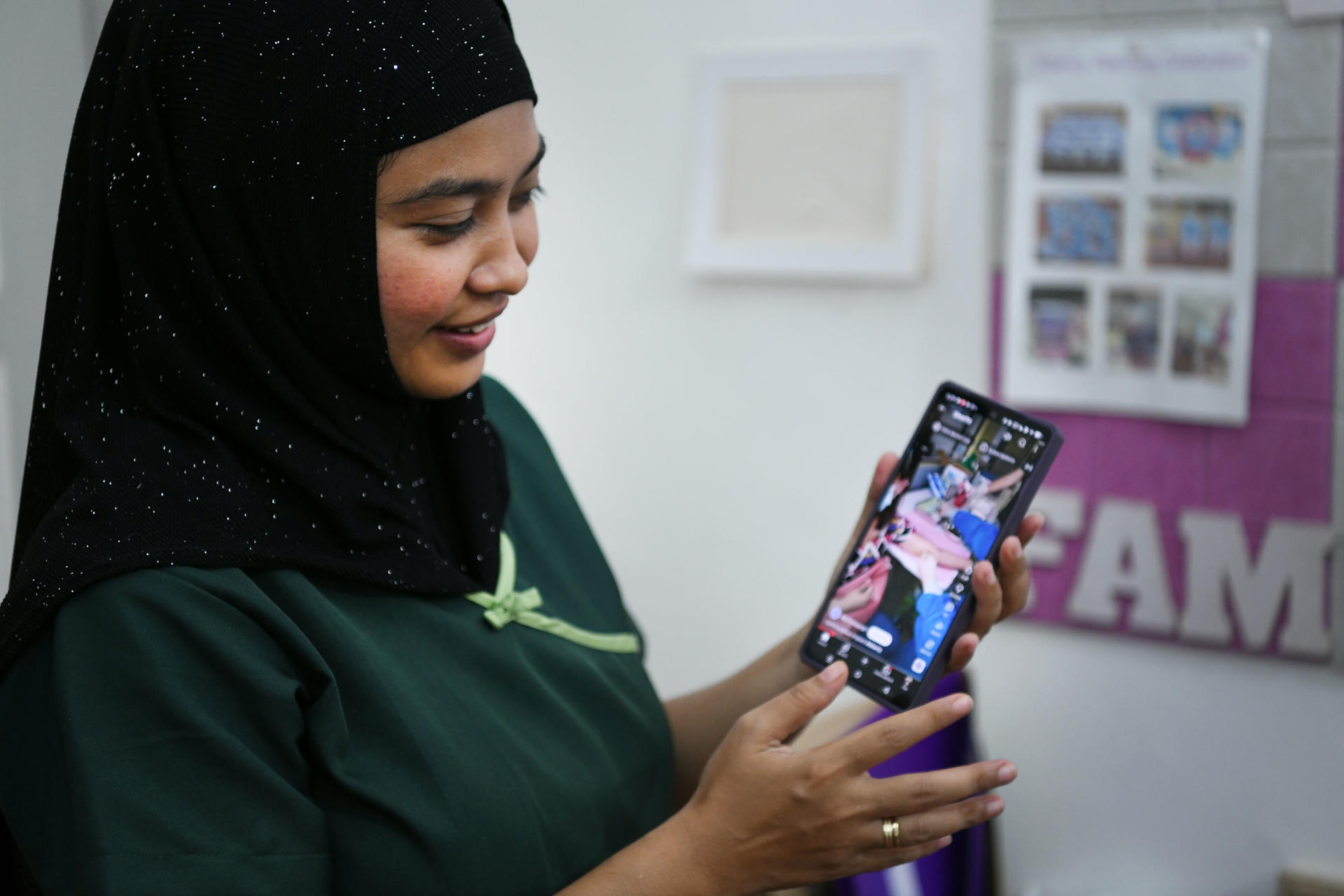 Woman in black hijab and green shirt, looking at a smartphone, holding a purple bottle.