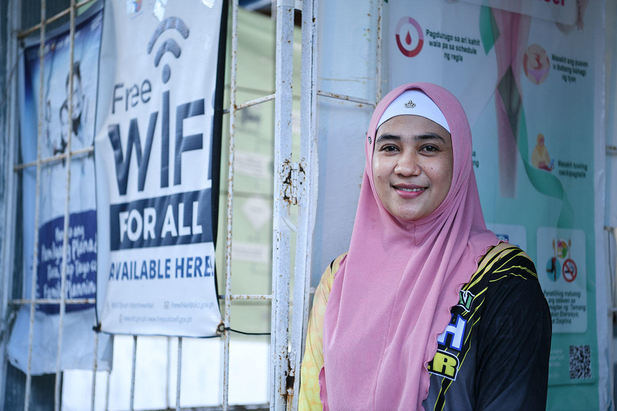 Young woman in hijab smiles beside a storefront with Free WiFi for all banners.