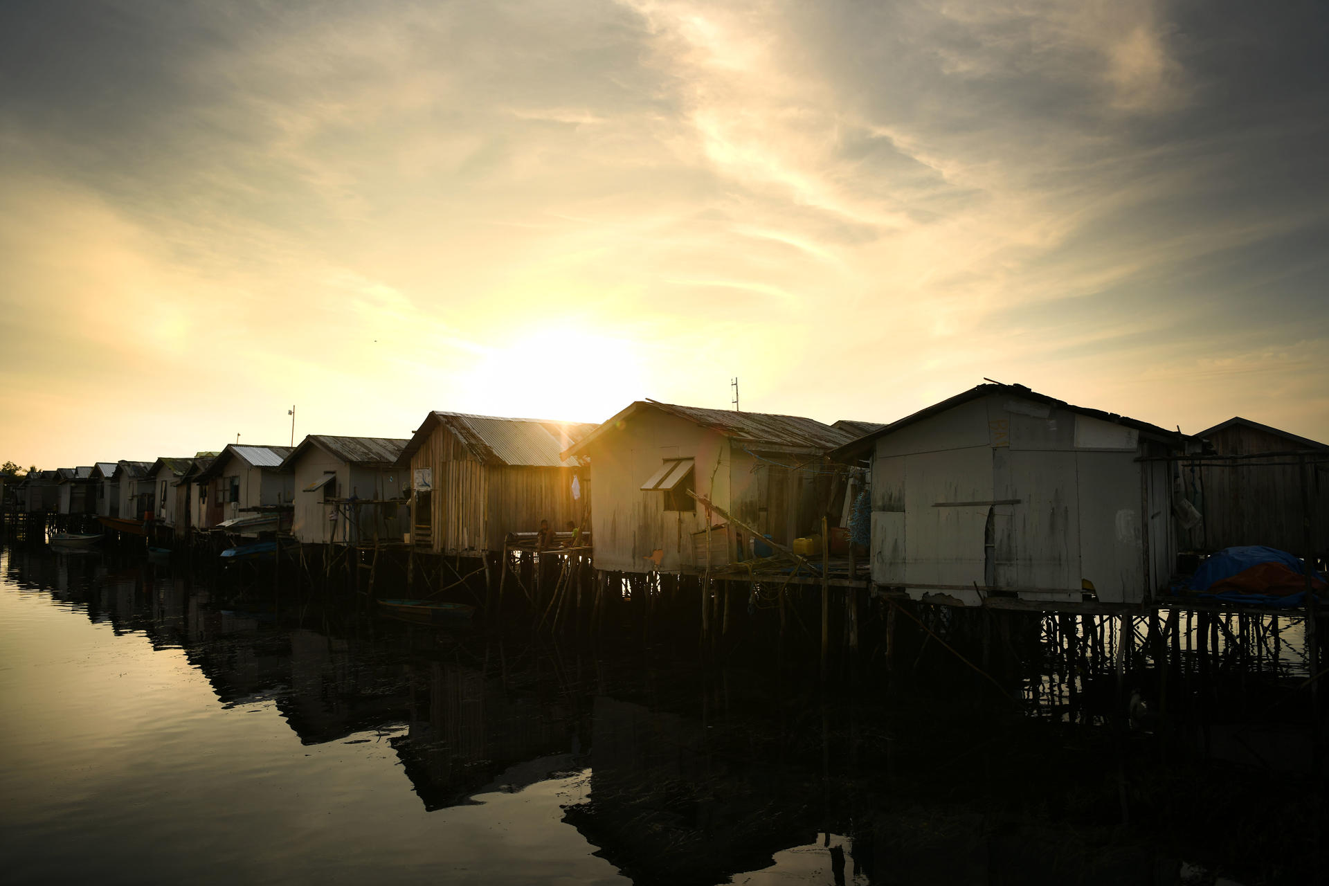 Row of stilt houses over calm water at sunset, glowing orange sky and reflections.
