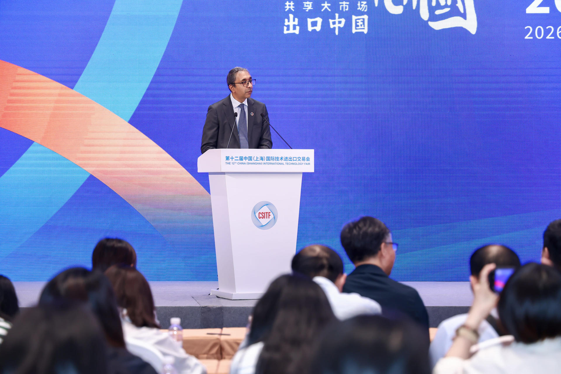 Photograph of a man in a suit giving a speech at a podium on stage with a blue backdrop.