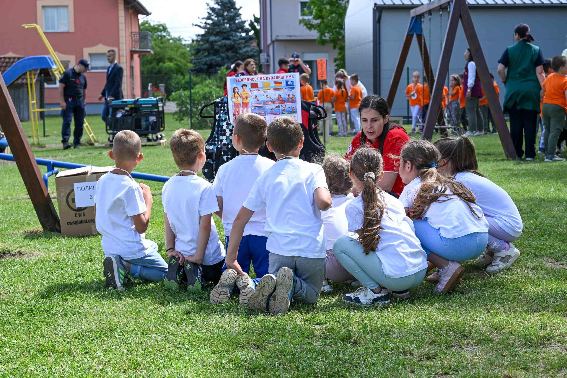 Children in white shirts sit in a circle on grass; playground equipment in background.