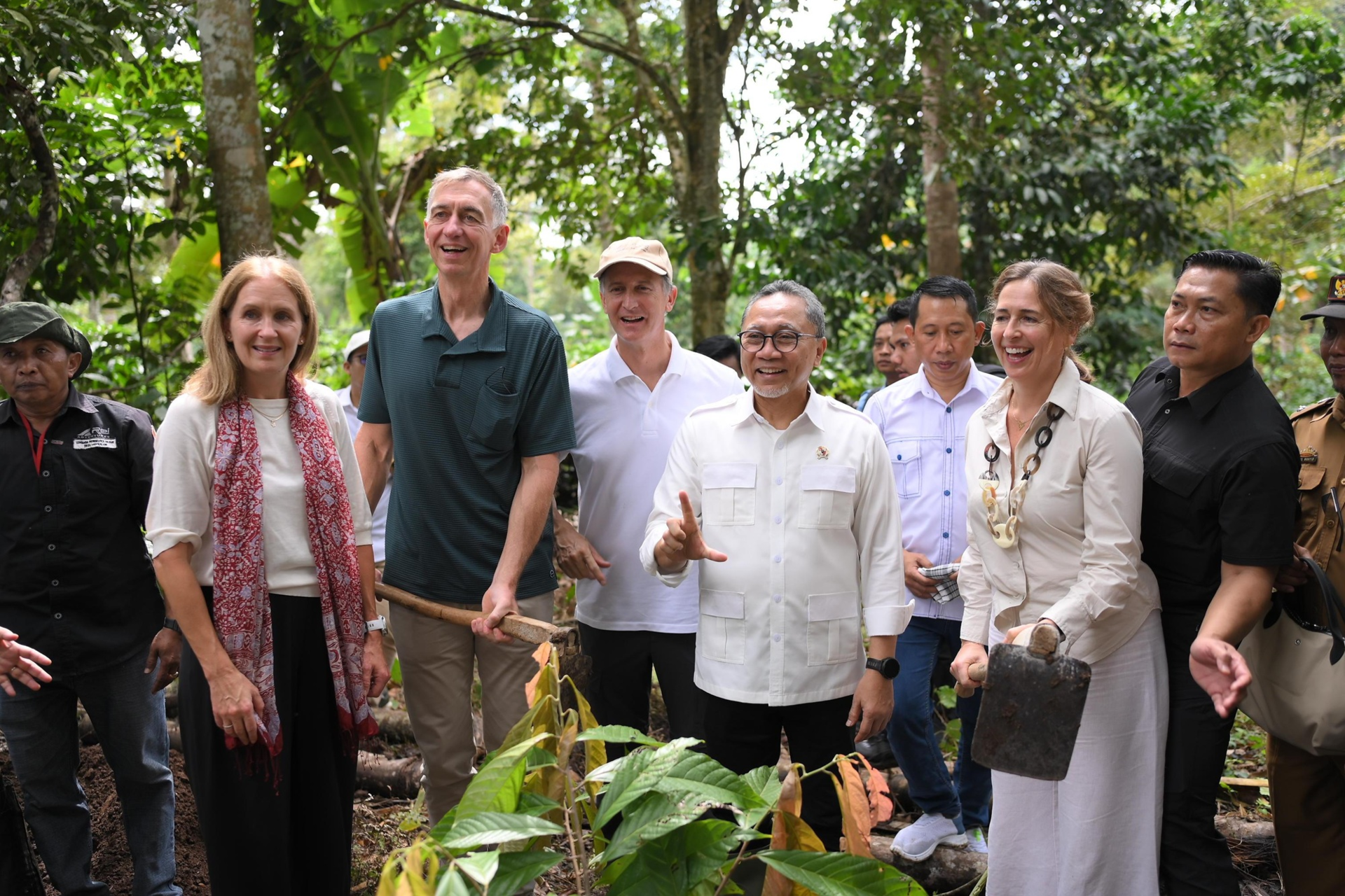 Diverse group of people smiling outdoors in a garden, gathered around a leafy plant.