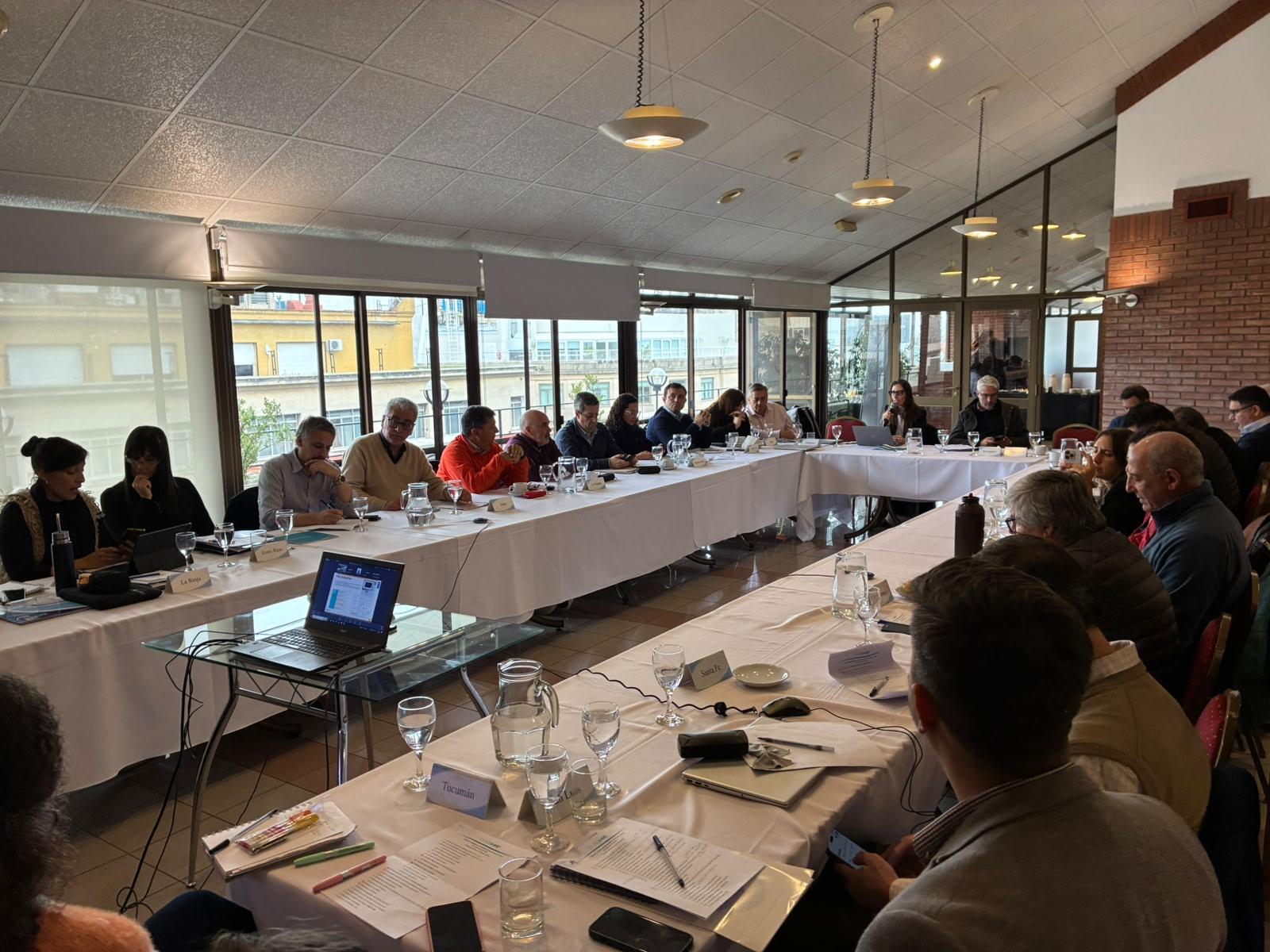 Large conference room with attendees around a long U-shaped table; laptops and papers.