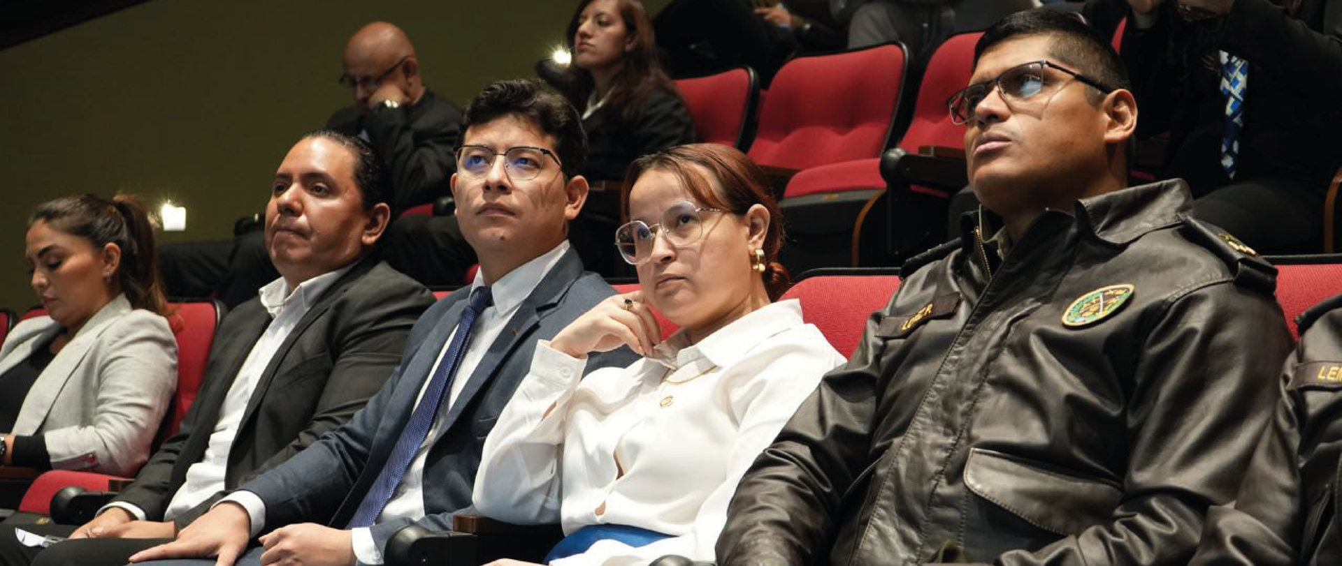 Photograph of an audience seated in red theater seats, faces blurred.