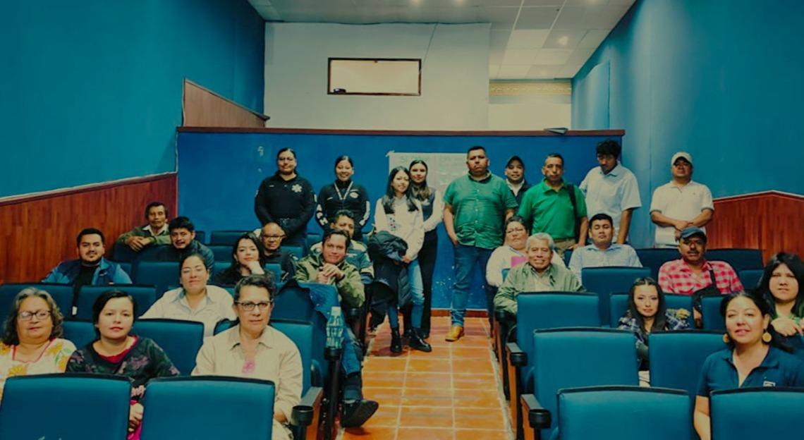 Group of people posing in a teal-walled classroom with blue seats and orange floor.