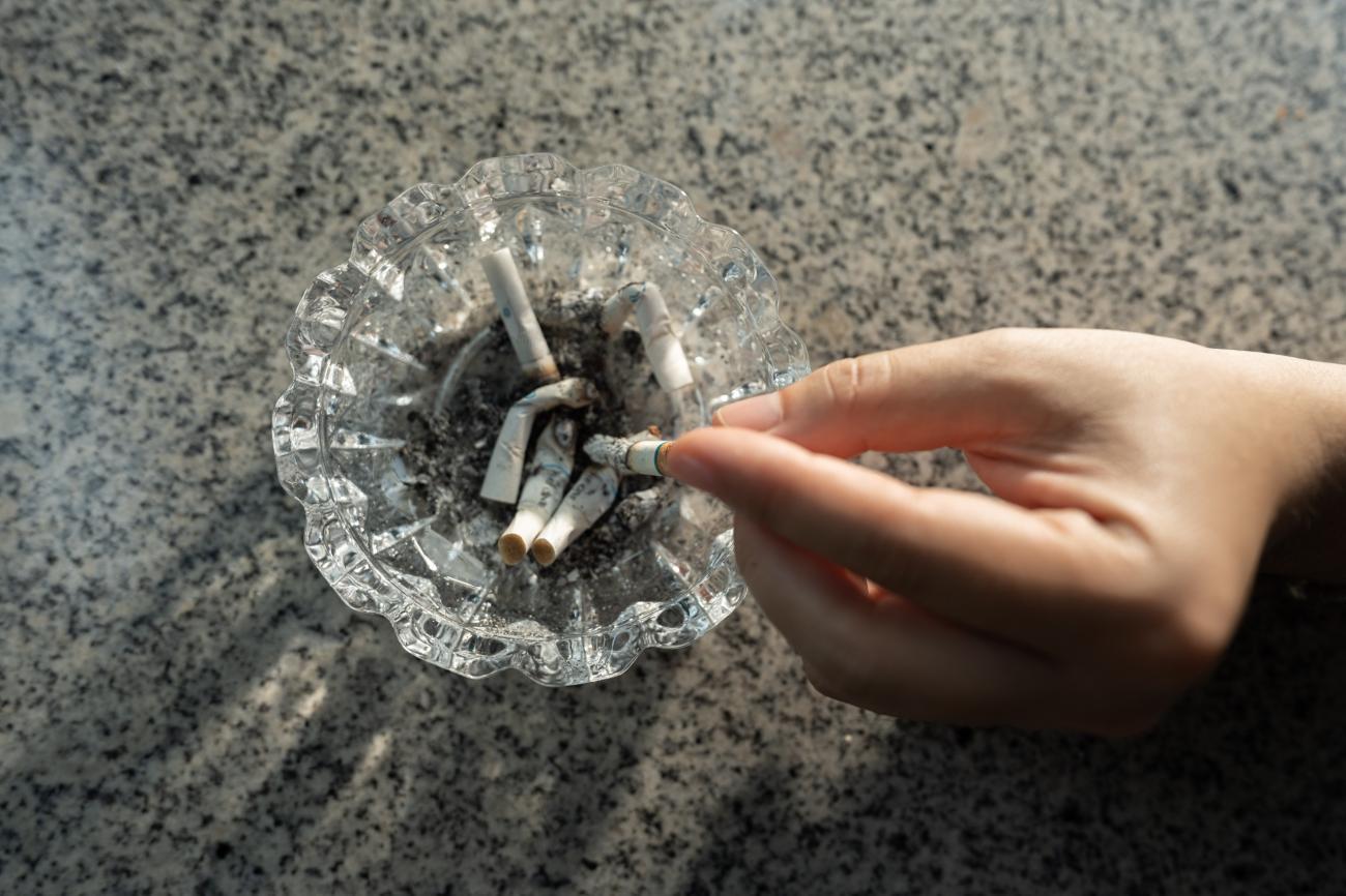 Hand placing a cigarette into a glass ashtray on a speckled countertop.