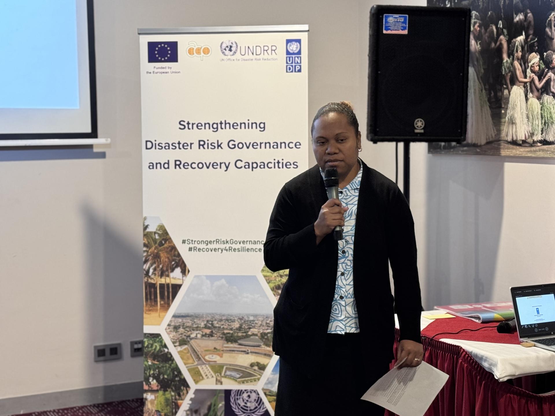 Speaker in dark blazer with microphone at a conference; disaster risk banners in the background.