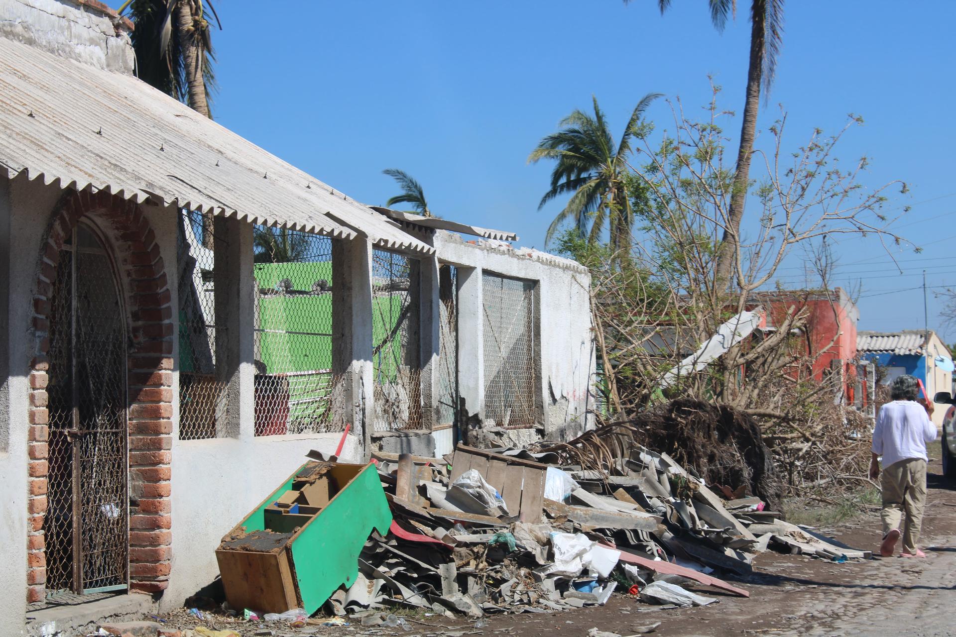 Rubble-strewn street with damaged houses, palm trees, and a lone passerby.