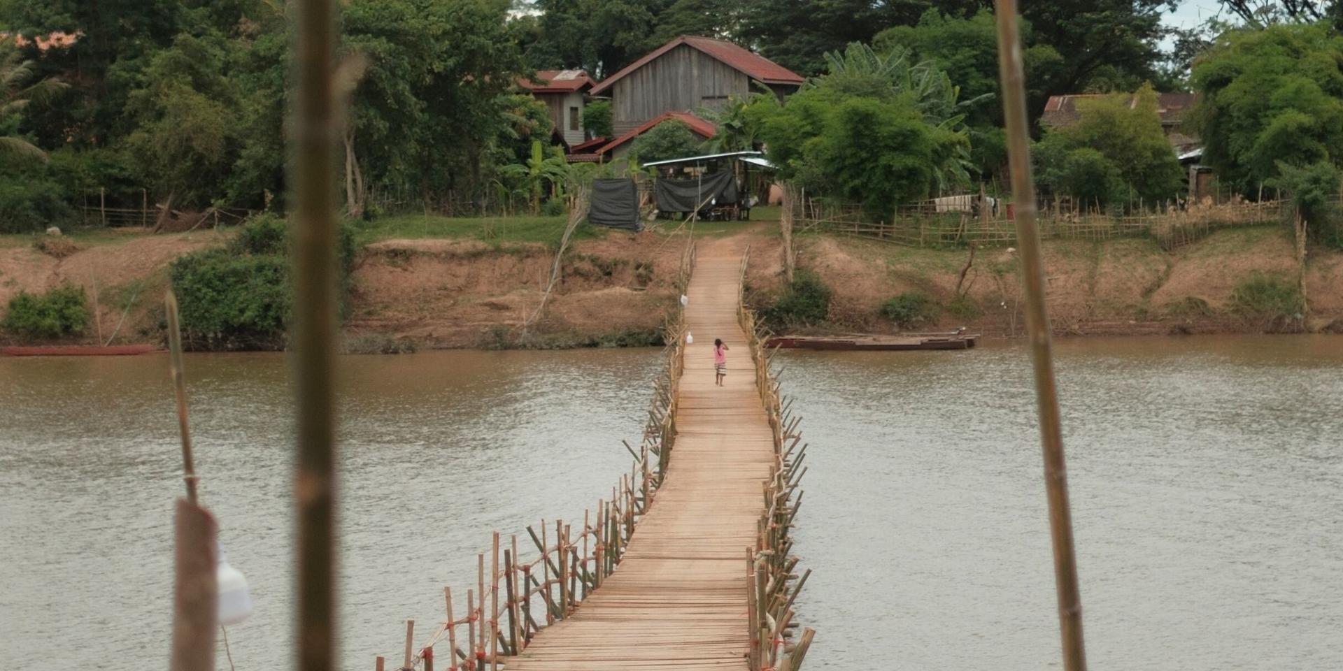 Narrow wooden footbridge across a river toward a hillside house among trees.