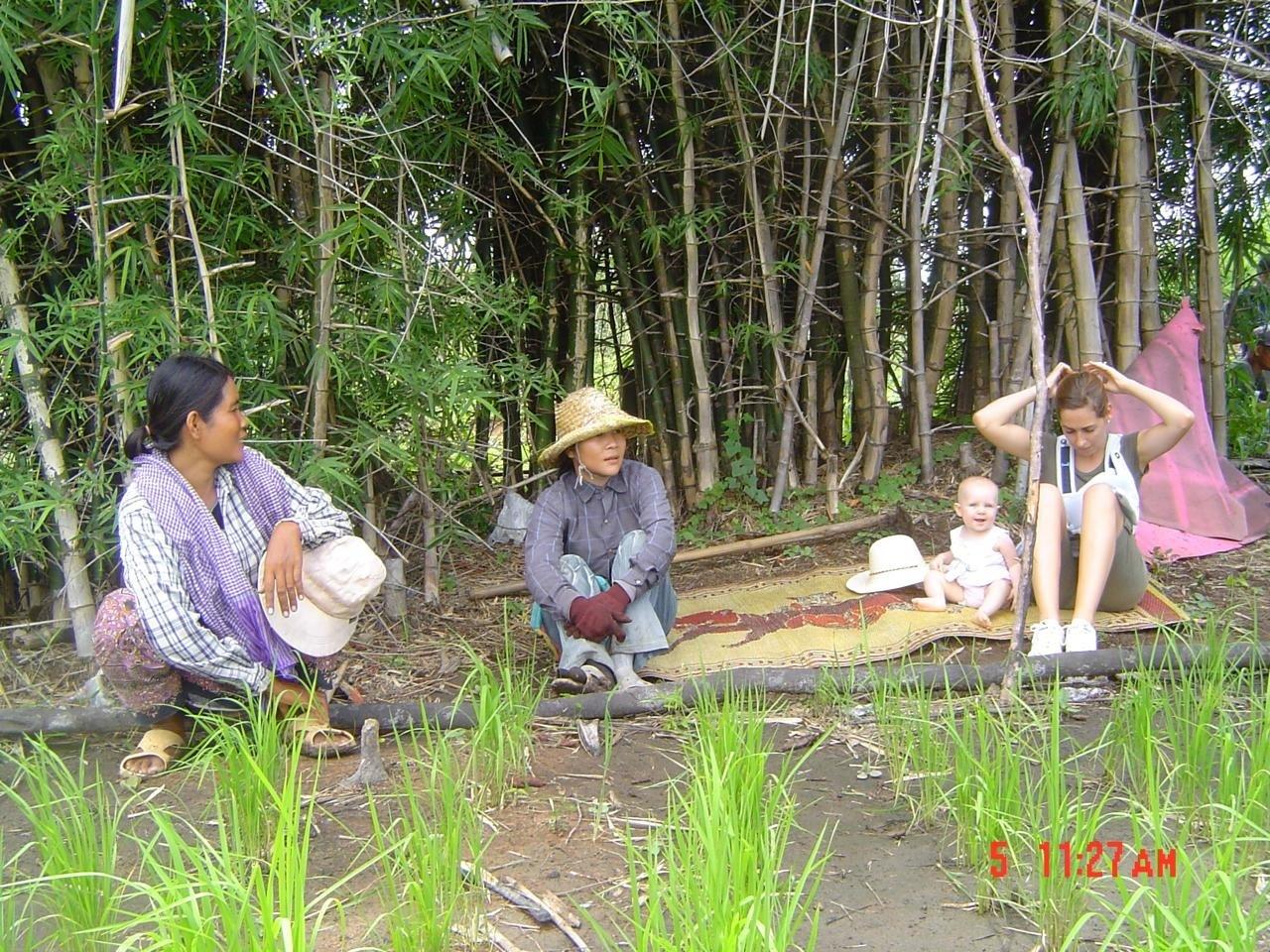 Family picnicking by a forest stream on mats; man with hat, child with pink umbrella.