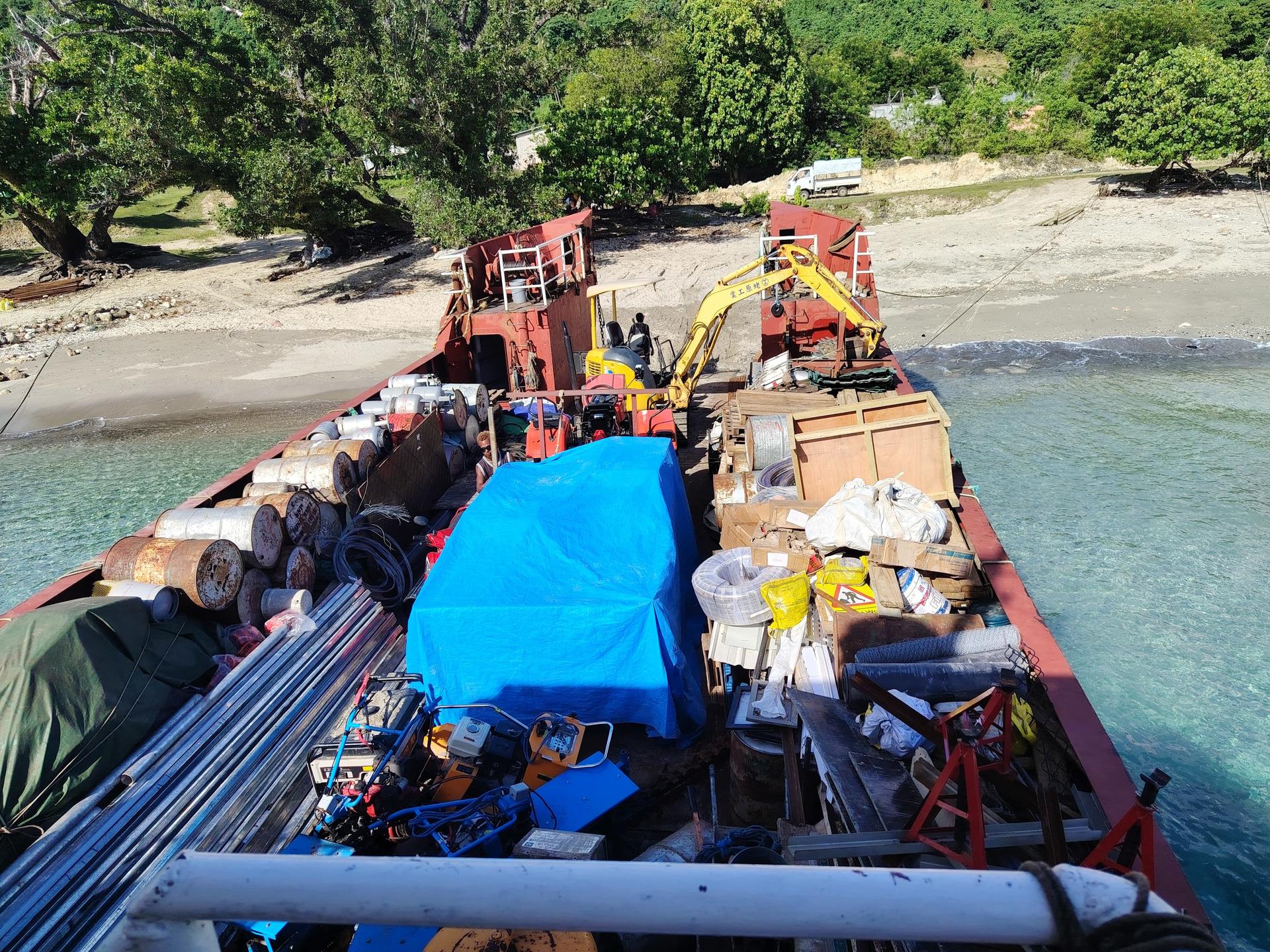 Photograph of a cargo barge on a river, with a blue tarp, crates, pipes, and crew aboard.