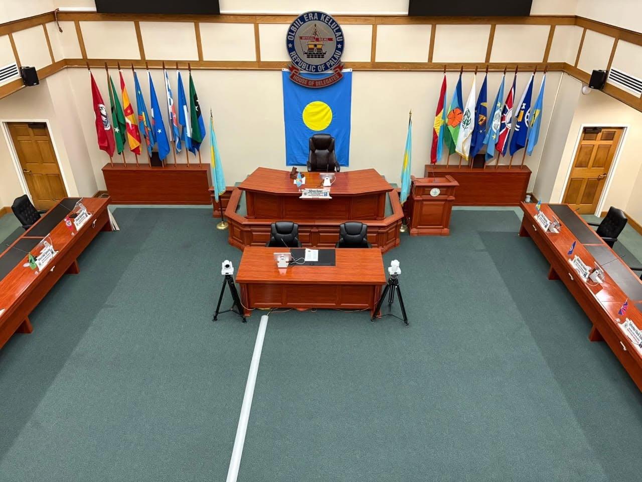 Empty council chamber with blue crest on wall behind a central dais and flags along the sides.