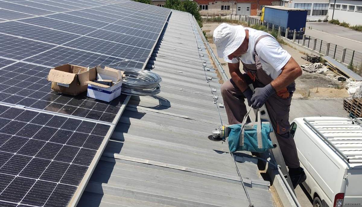 Photograph of a construction worker on a roof beside solar panels, wearing a hard hat.