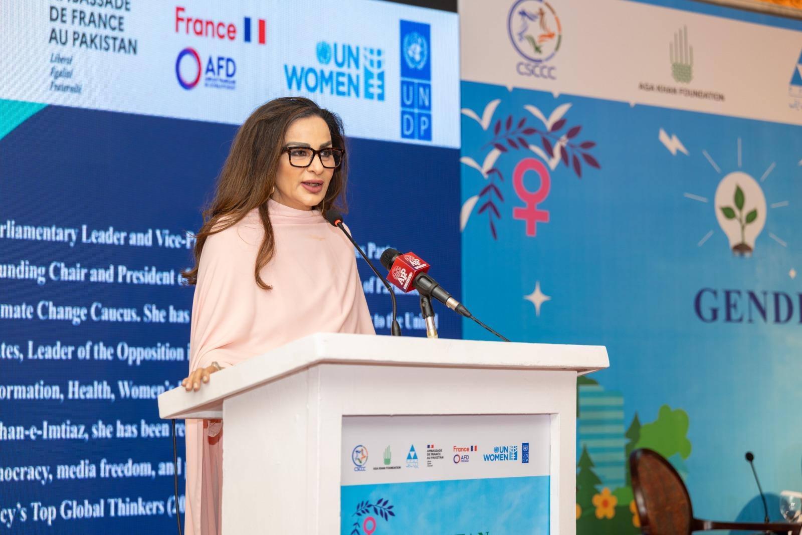 Woman in pink top speaks at a podium during a conference, blue banners with logos behind.