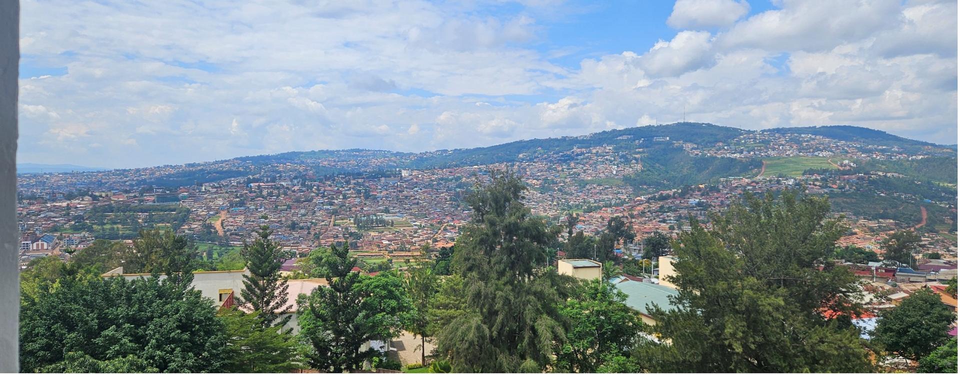 Panoramic view of a green valley with trees, houses, and distant hills under a blue sky.